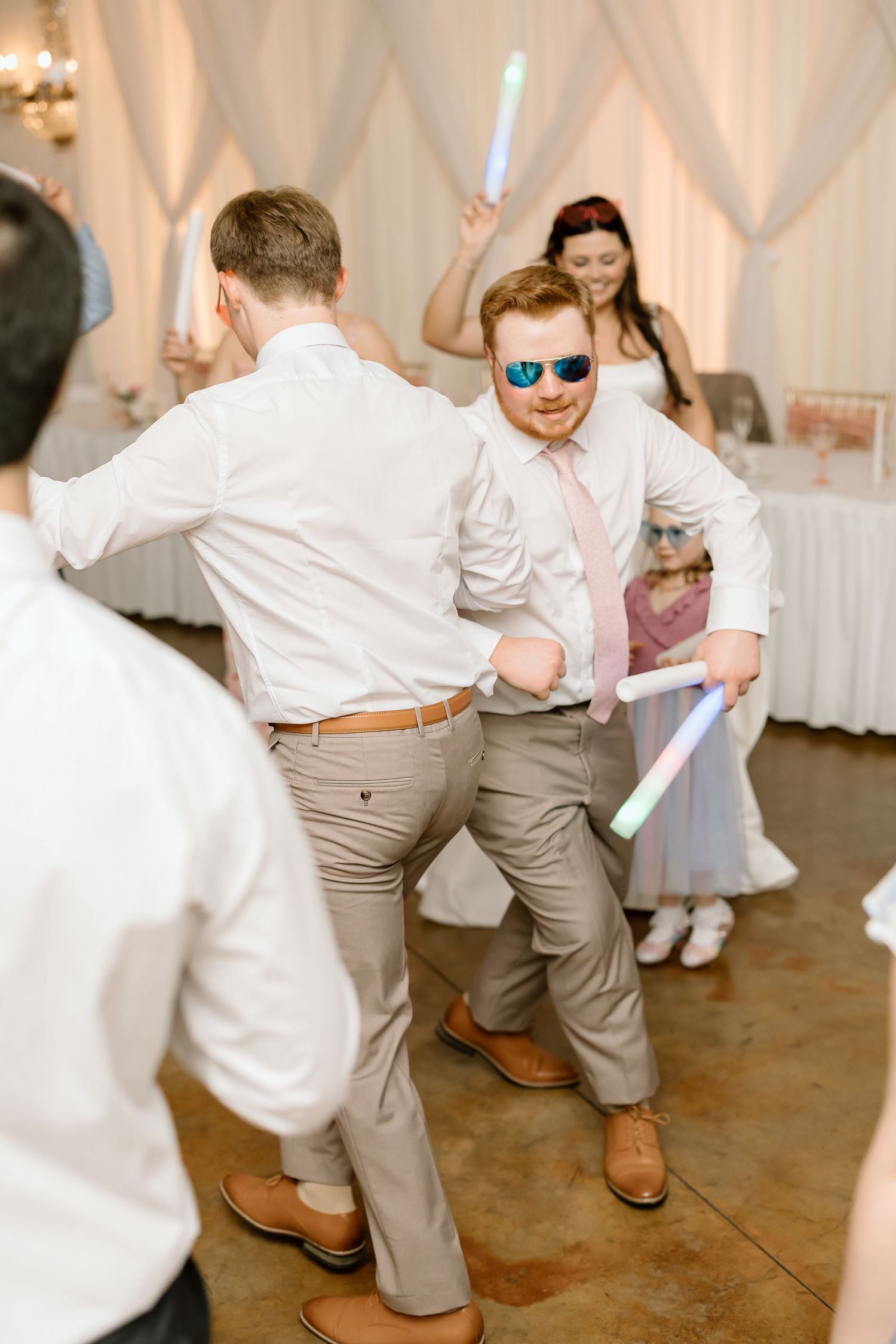 Wedding guests dance and celebrate on the dance floor at a reception with white decor and festive atmosphere.