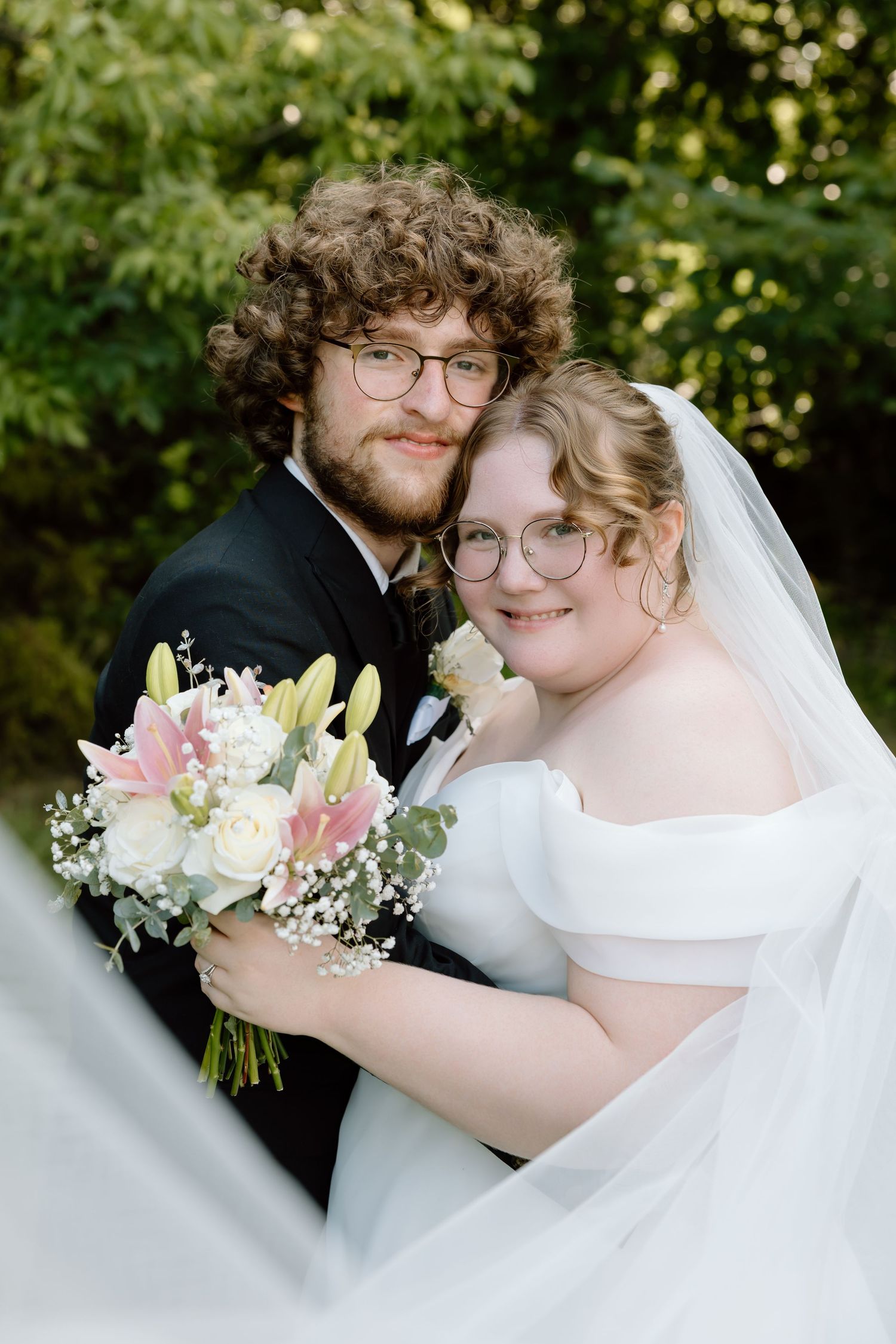 A smiling bride and groom in wedding attire pose outdoors holding a bouquet of pink and white flowers.