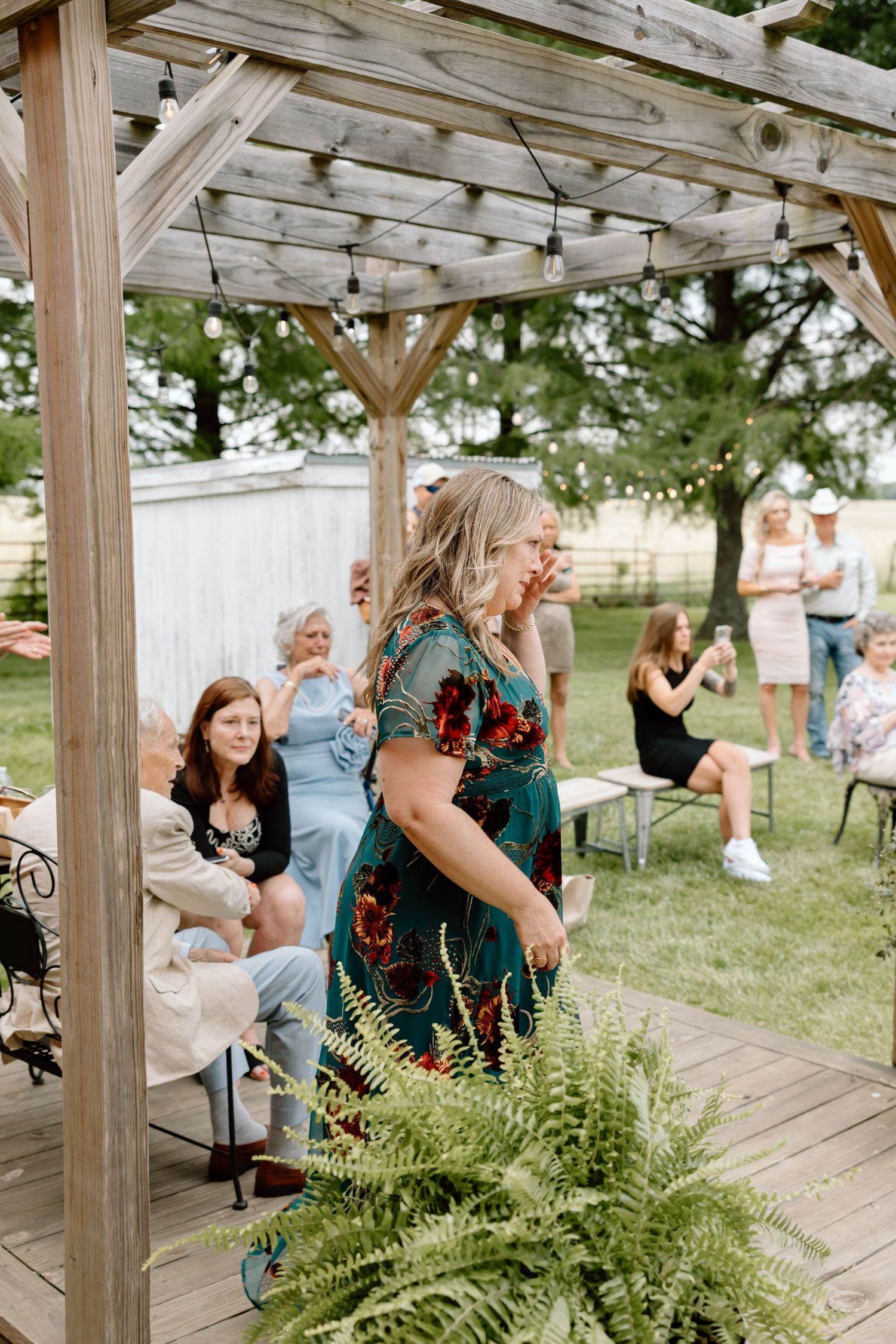 Women gather under a rustic wooden pergola with string lights at an outdoor farm party on a sunny day.