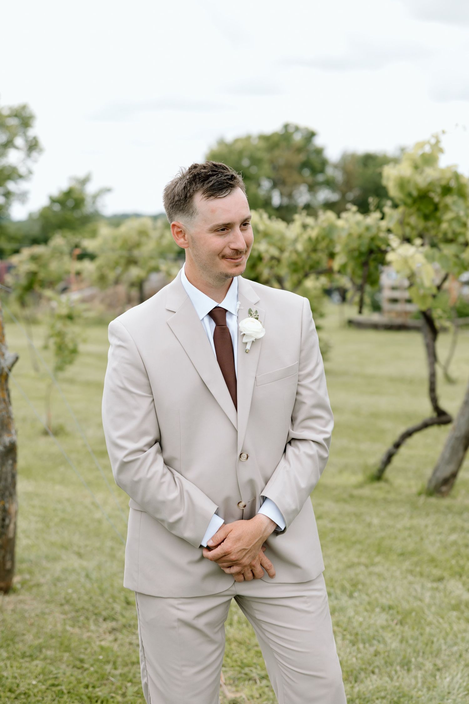 Groom in beige suit with brown tie and white boutonniere smiling outdoors at a garden wedding venue.