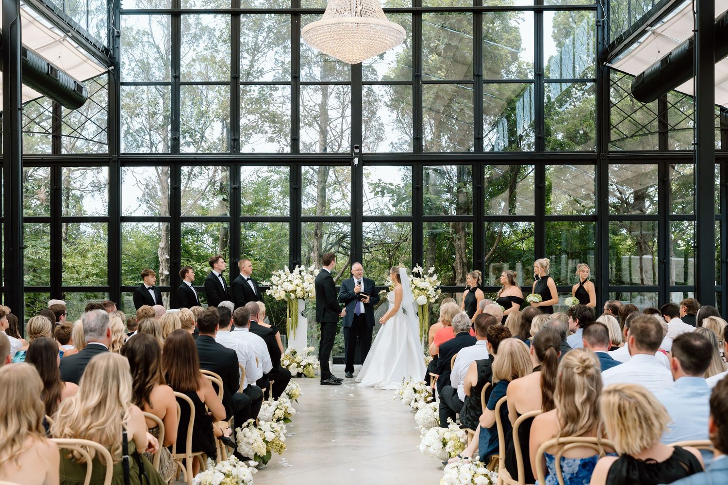 Elegant wedding ceremony in a glass greenhouse venue with white floral arrangements and guests seated in navy chairs.