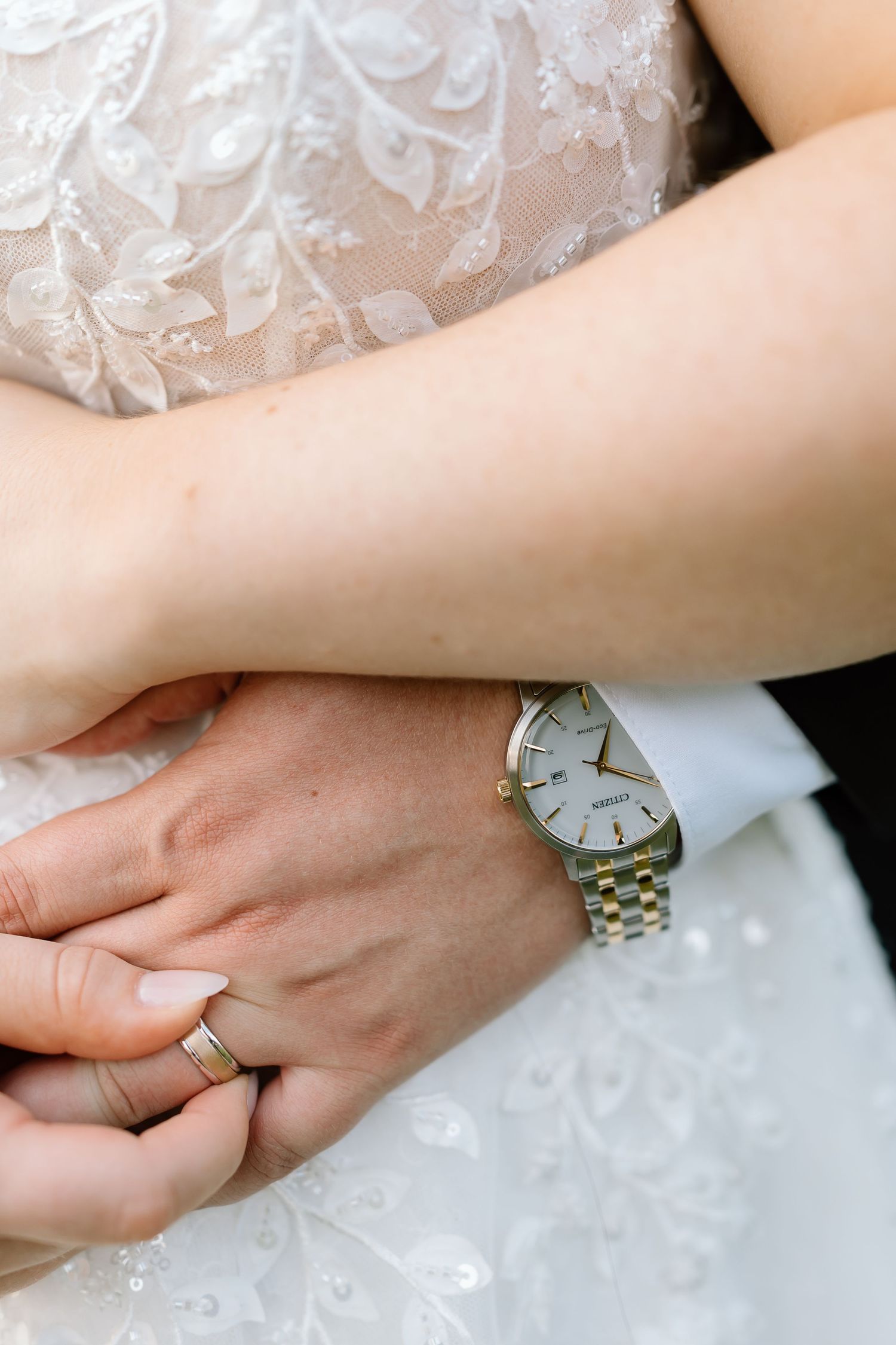 Close-up view of hands wearing engagement ring and watch against white lace fabric background.