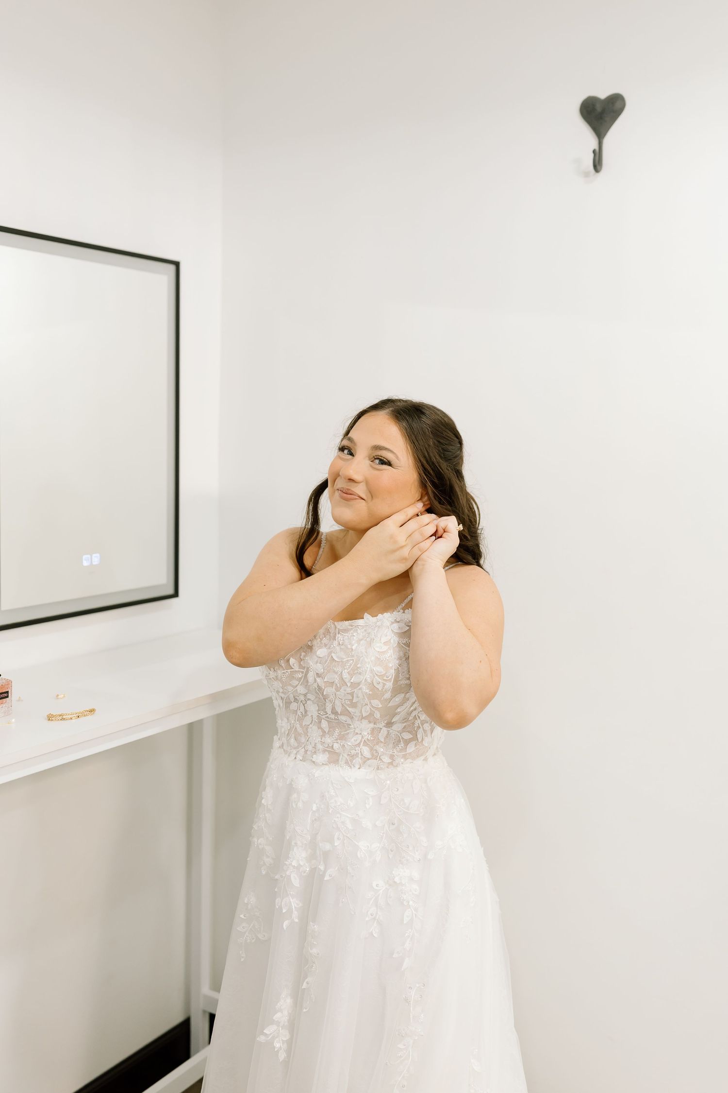 A bride in a white lace wedding dress adjusts her hair while getting ready in a bright room.
