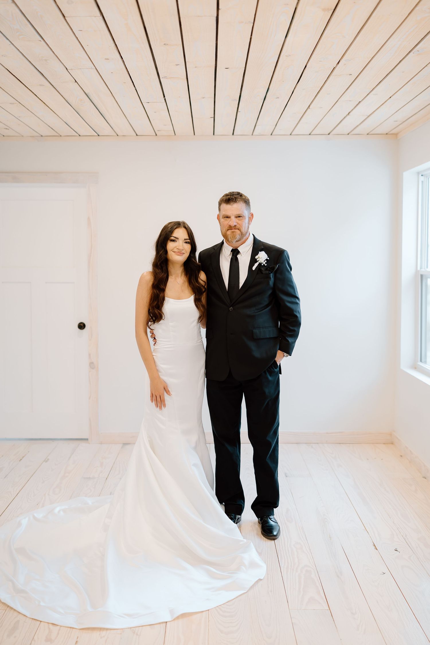 A couple poses in a minimalist white room with wooden floors and a flowing white wedding gown train.