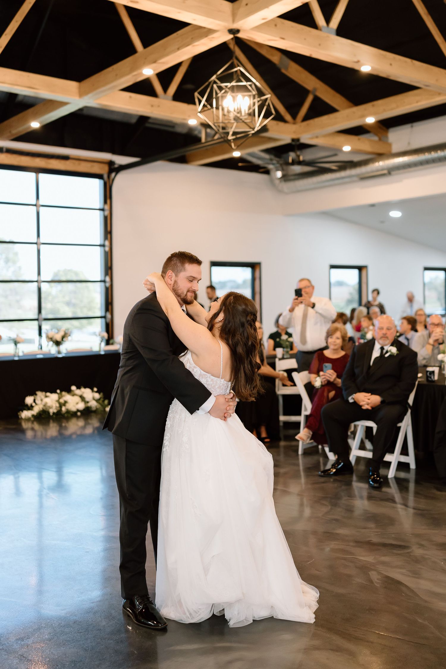 A couple shares a romantic dip during their first dance at a modern wedding reception venue with exposed beams.