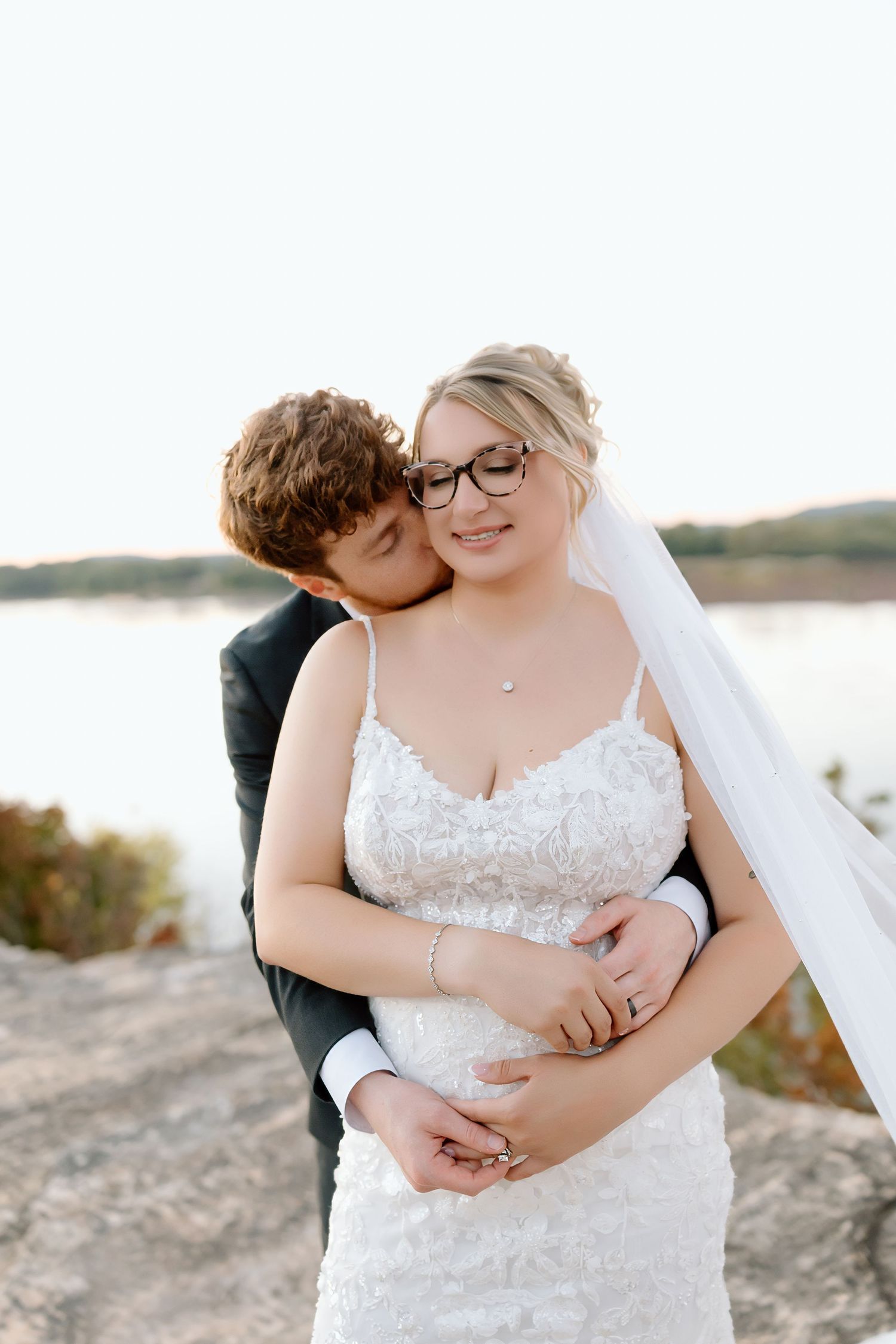 A romantic wedding couple embraces on a rocky beach at sunset with a water view in the background.