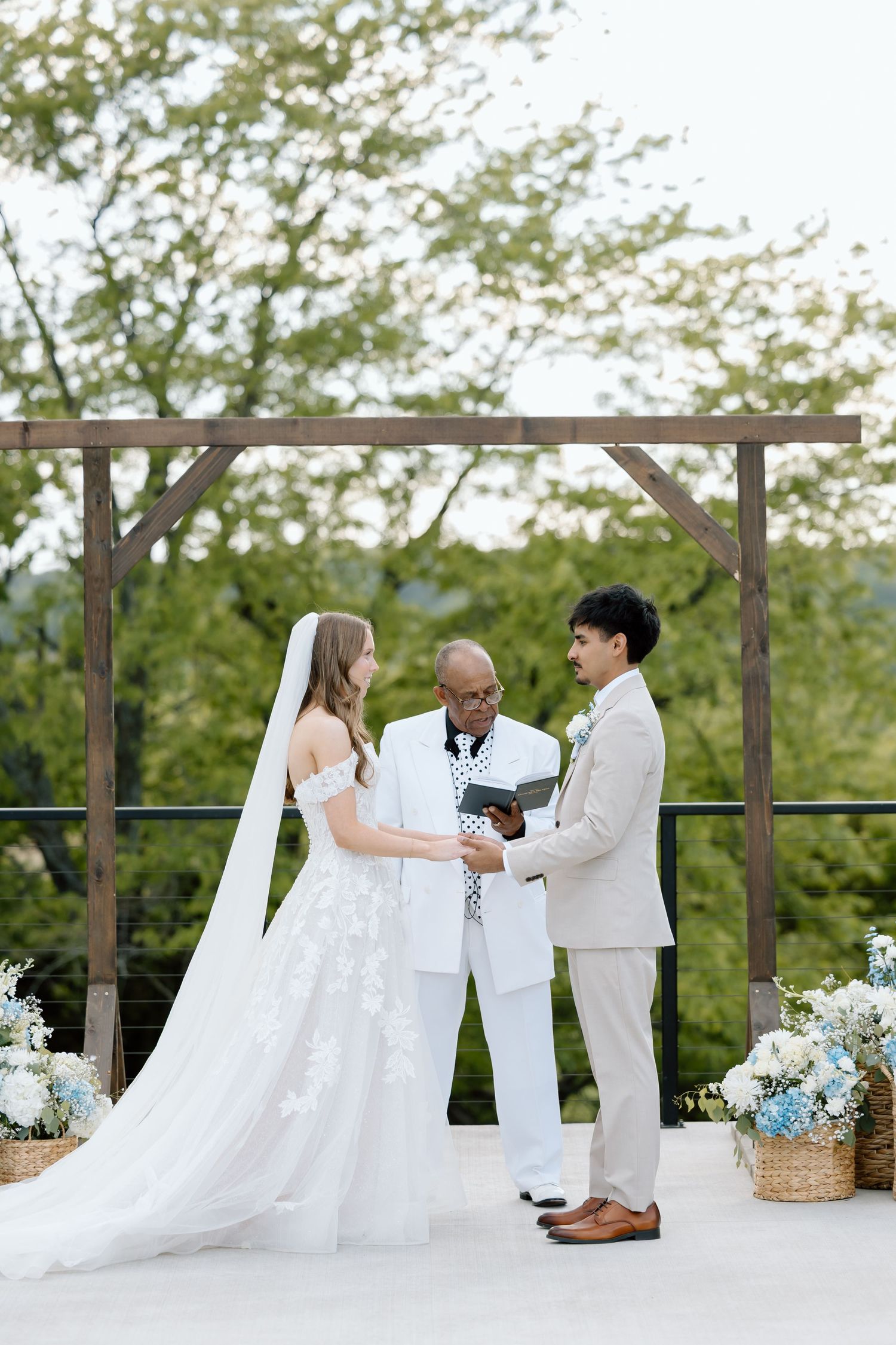 Outdoor wedding ceremony with bride in lace gown, groom in tan suit, and officiant under wooden arch with floral arrangements.