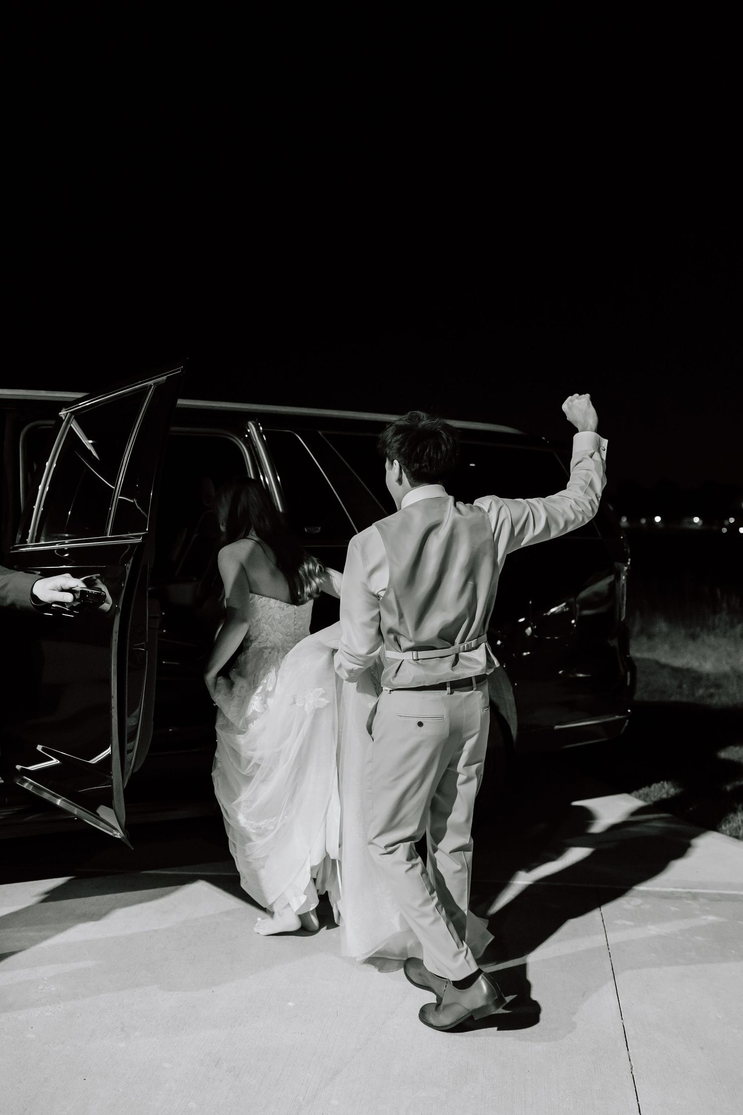 Black and white photo of a bride and groom celebrating as they enter a limousine at night after their wedding.