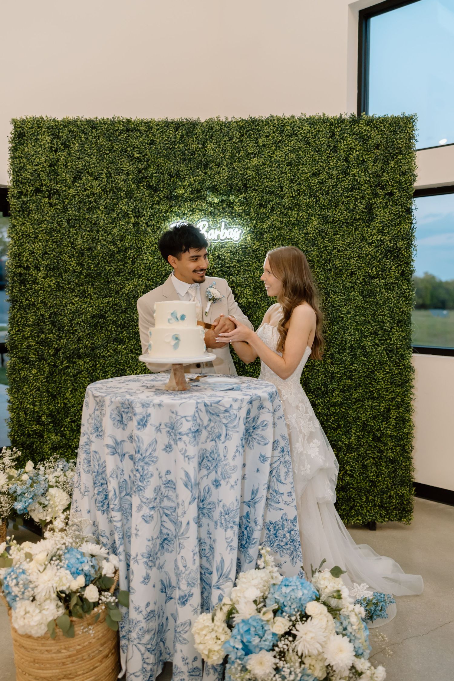 Bride and groom cutting a white and blue wedding cake at a floral table with a green hedge backdrop.