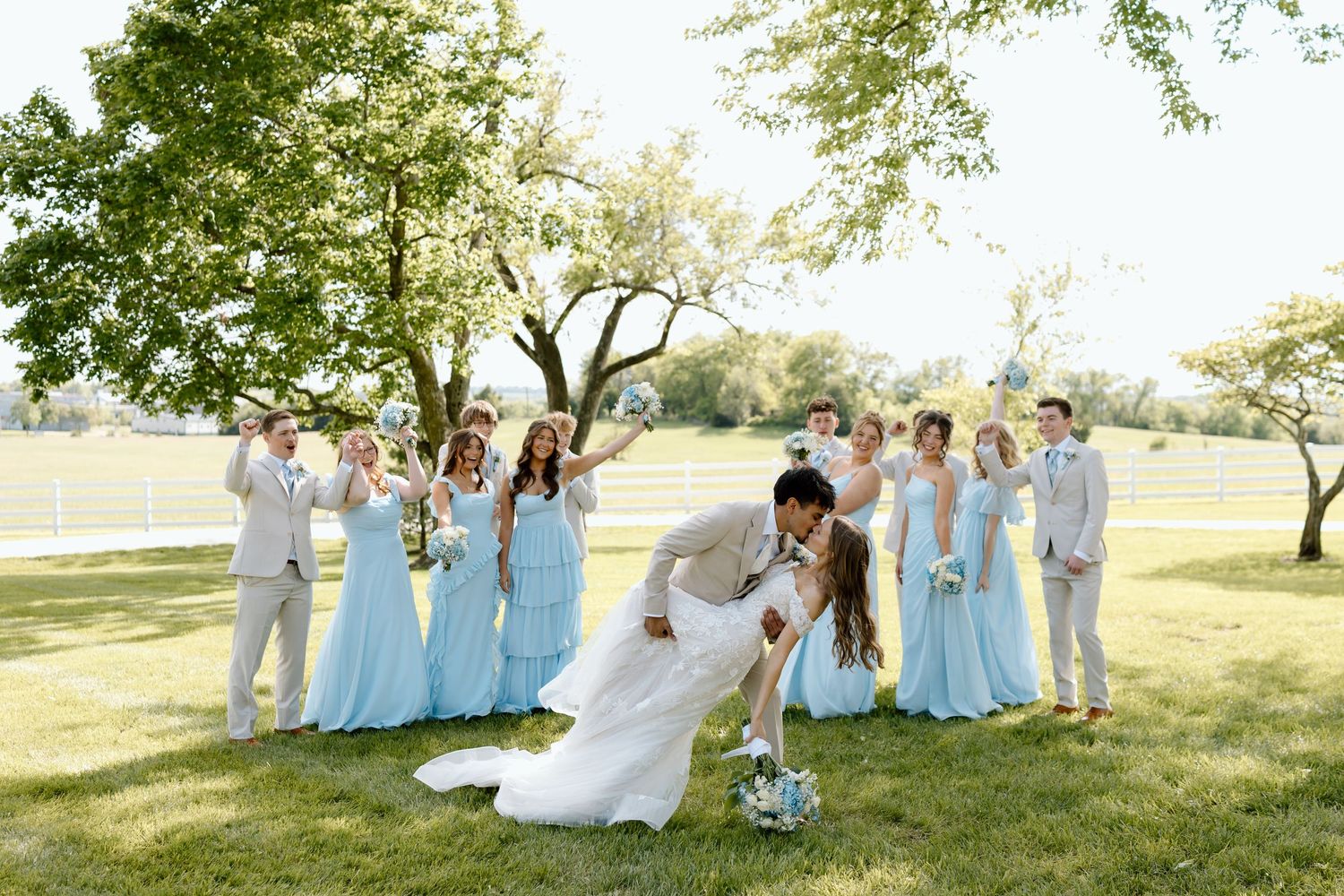 Wedding party poses outdoors as bride and groom share a romantic dip kiss on lush green lawn with bridesmaids in light blue gowns.