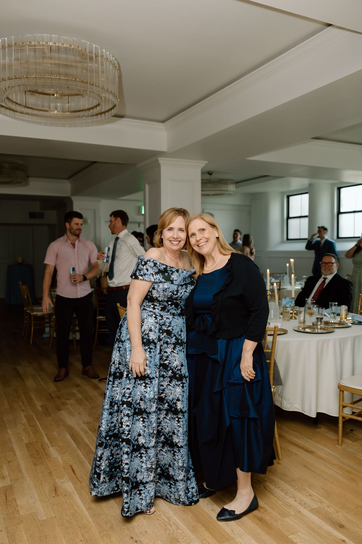 Two women smile together at an elegant wedding reception venue with chandelier lighting and candlelit tables.