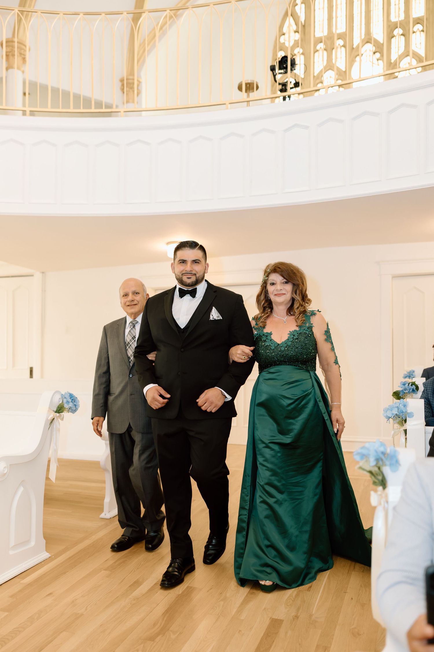Groom in black tuxedo walks down wedding aisle escorted by parents, woman in emerald green gown.