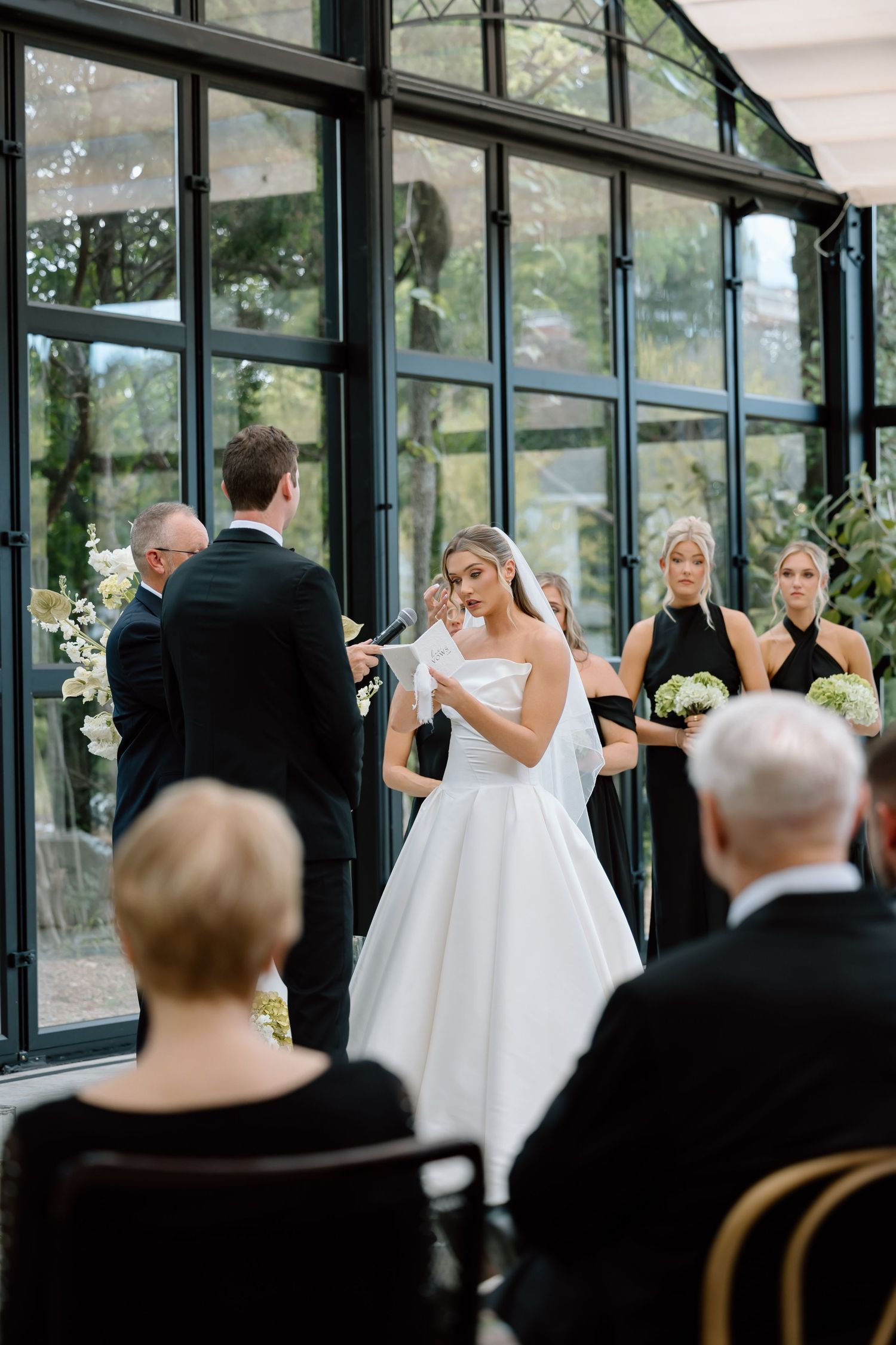 Bride reads vows in elegant white gown at greenhouse wedding ceremony with bridesmaids in black dresses.