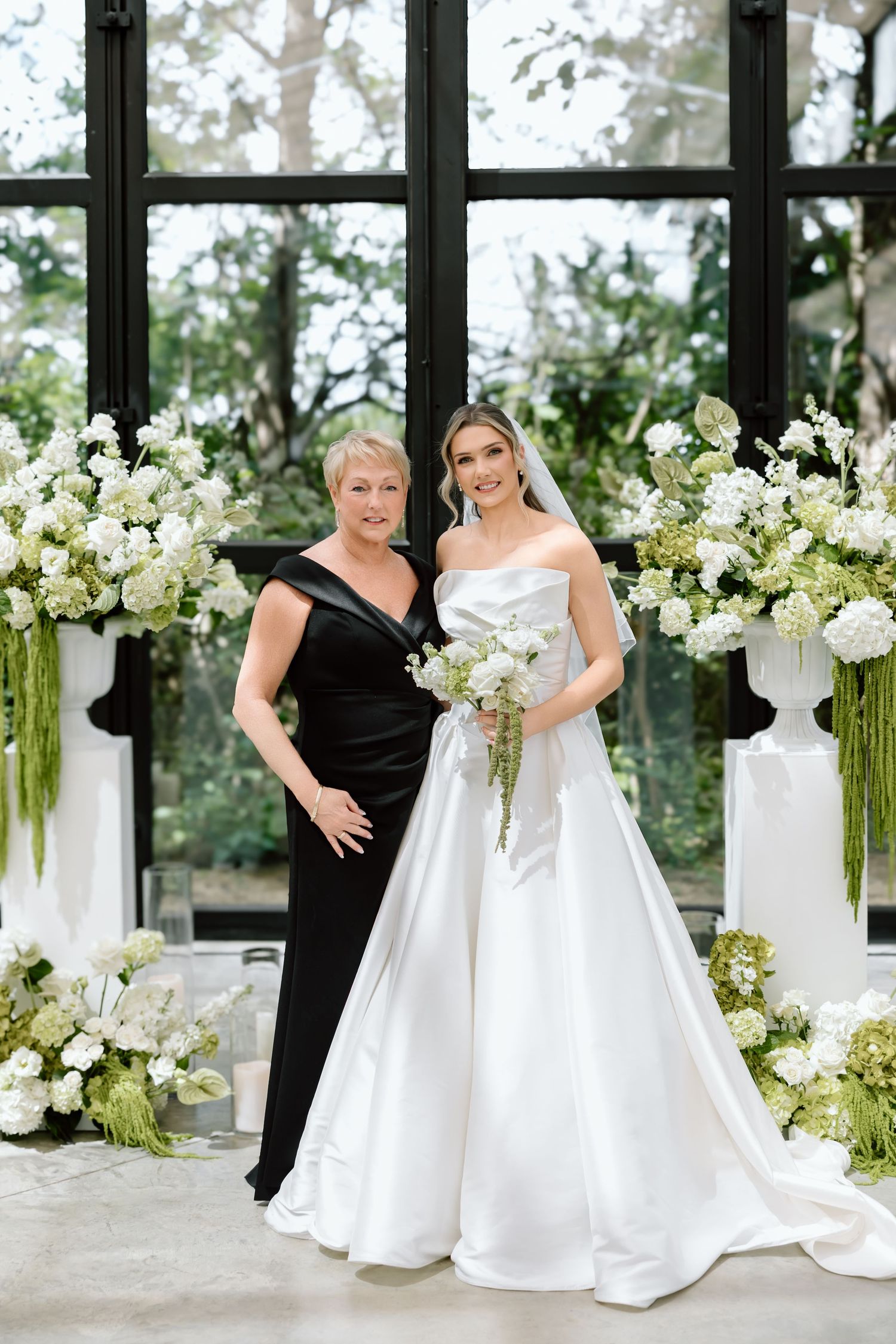 Bride in white strapless gown holding bouquet poses with woman in black dress among white floral arrangements.