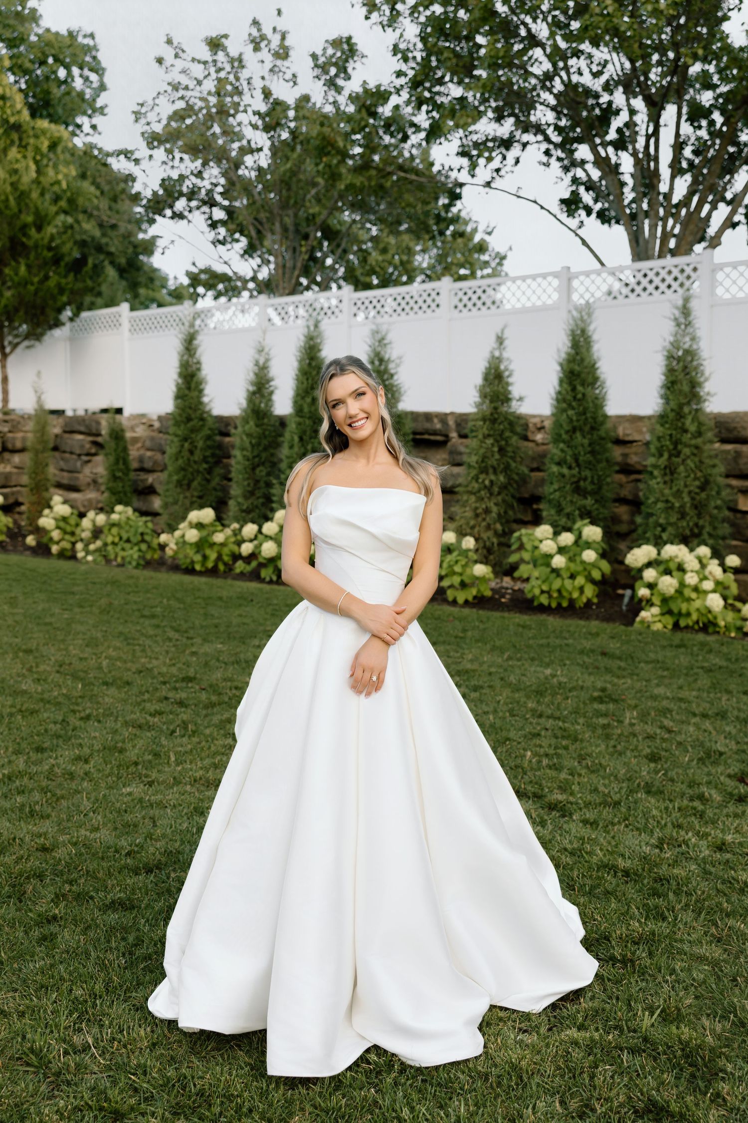 Bride in a strapless white ballgown standing on green lawn with hydrangeas and evergreen trees in background.