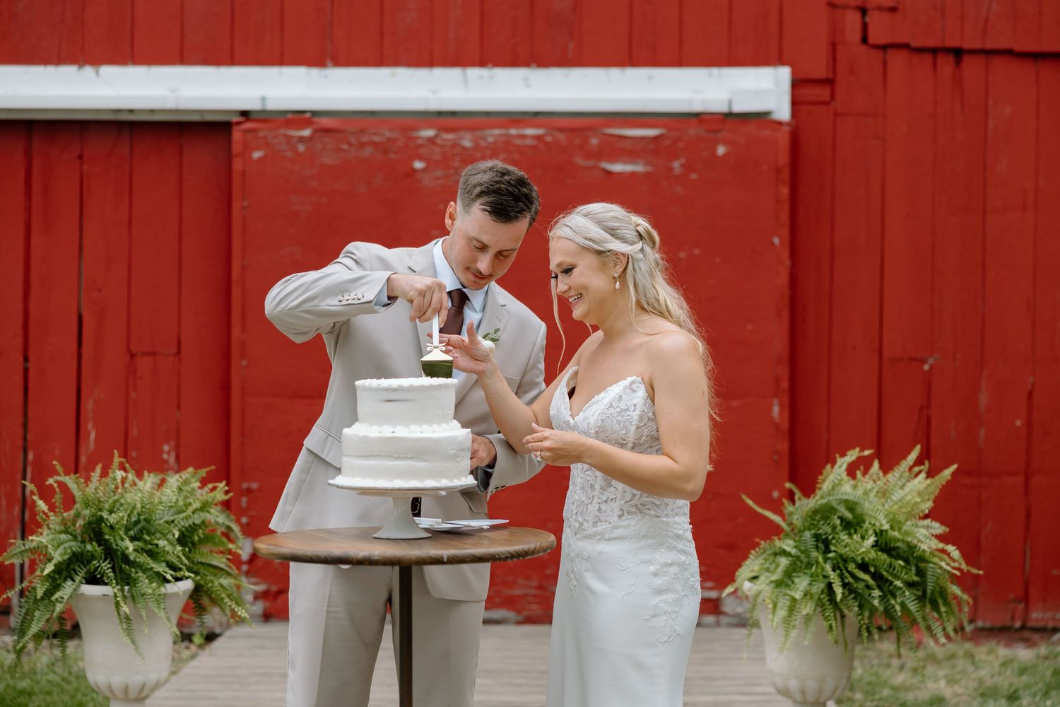 Bride and groom cutting white wedding cake outdoors in front of a red barn, surrounded by lush ferns.