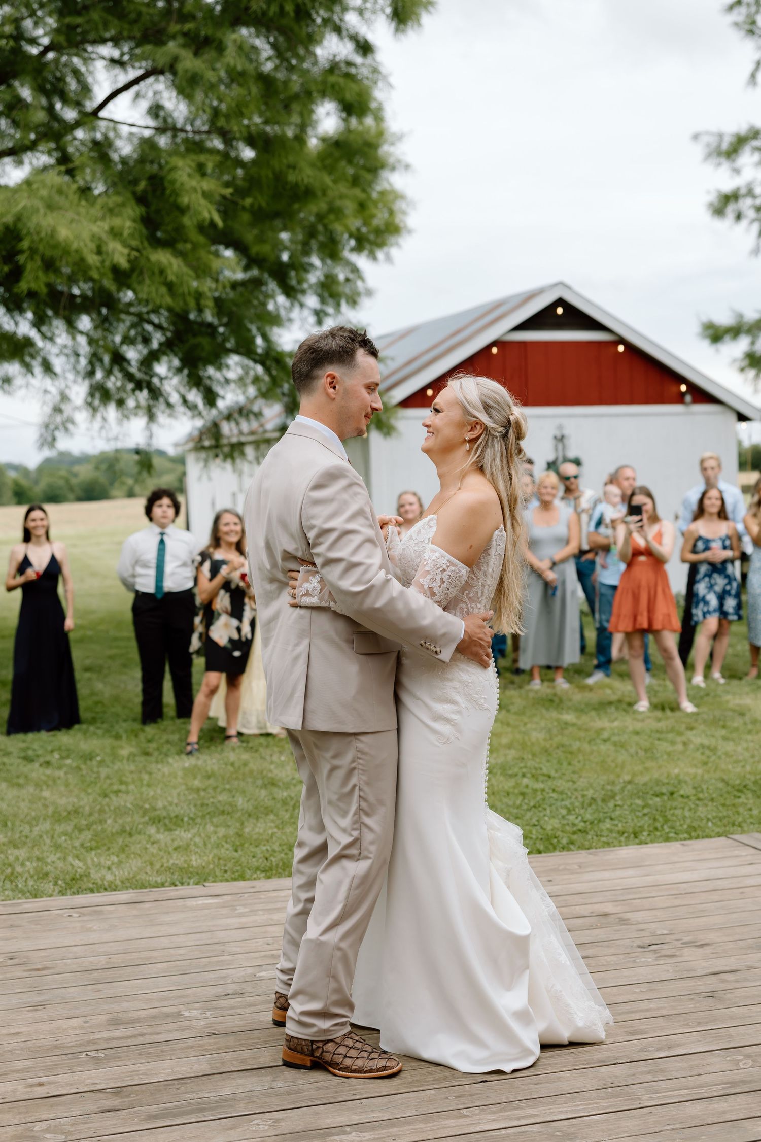 Bride and groom share first dance on outdoor wooden deck at rustic barn wedding venue with guests watching.