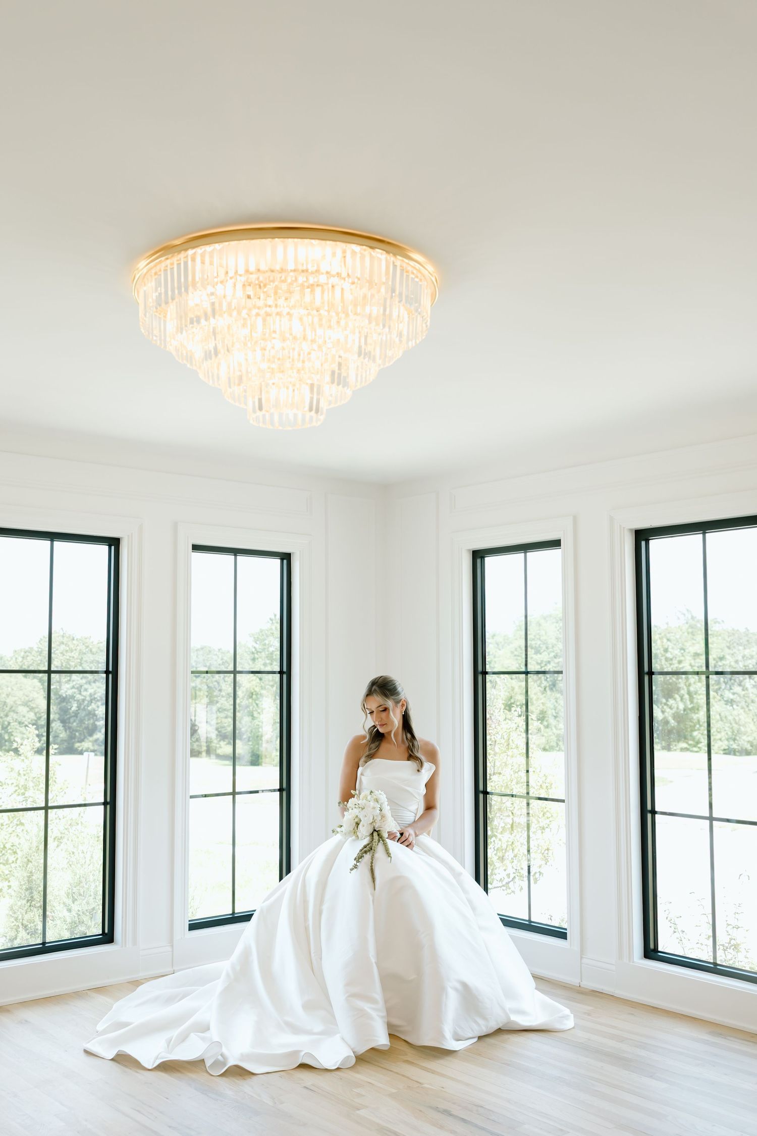 Bride in a white ballgown holding a bouquet stands in a bright room with black-framed windows and a crystal chandelier.
