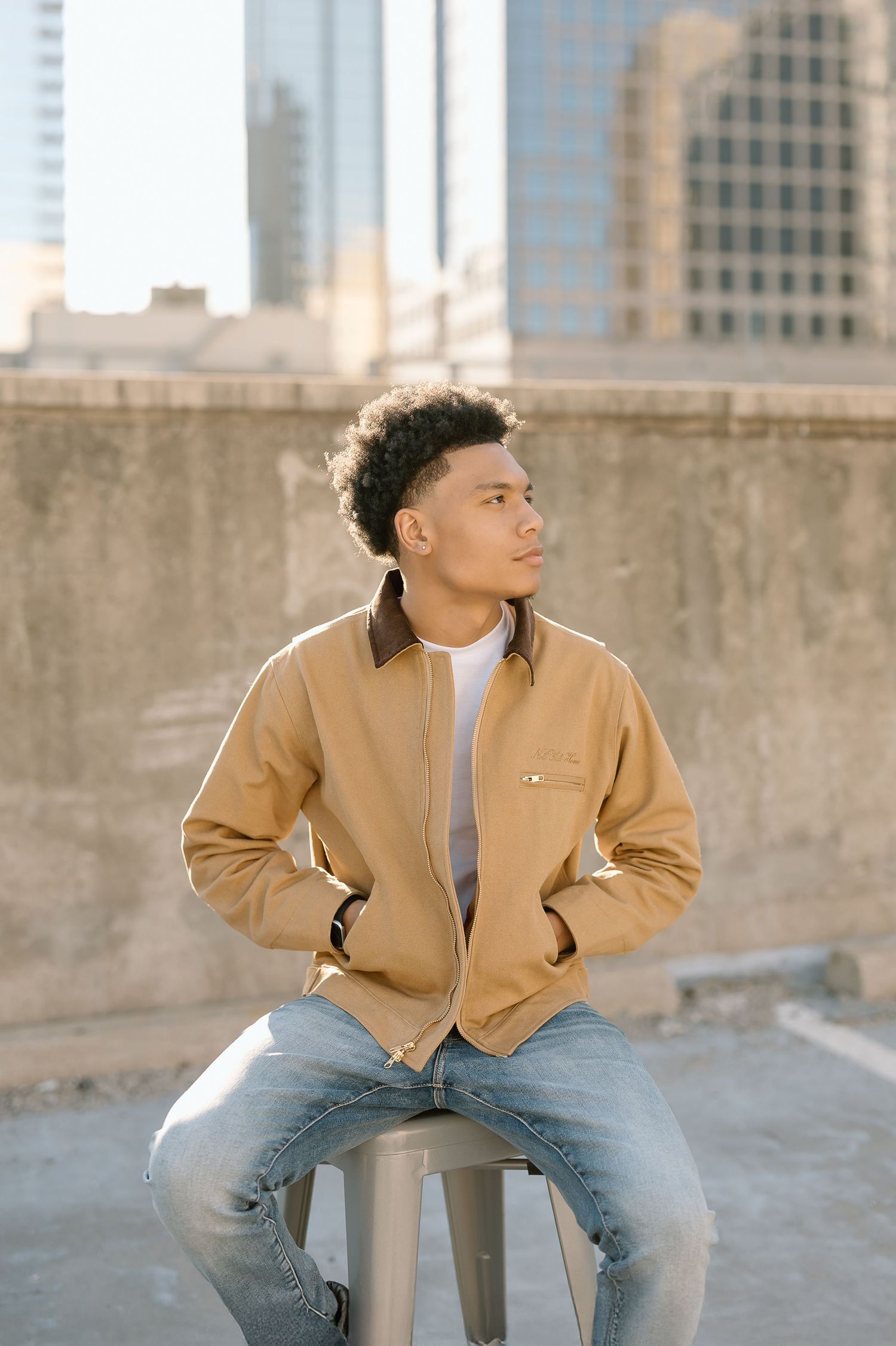 Person in tan jacket sits on stool with downtown skyline in background.