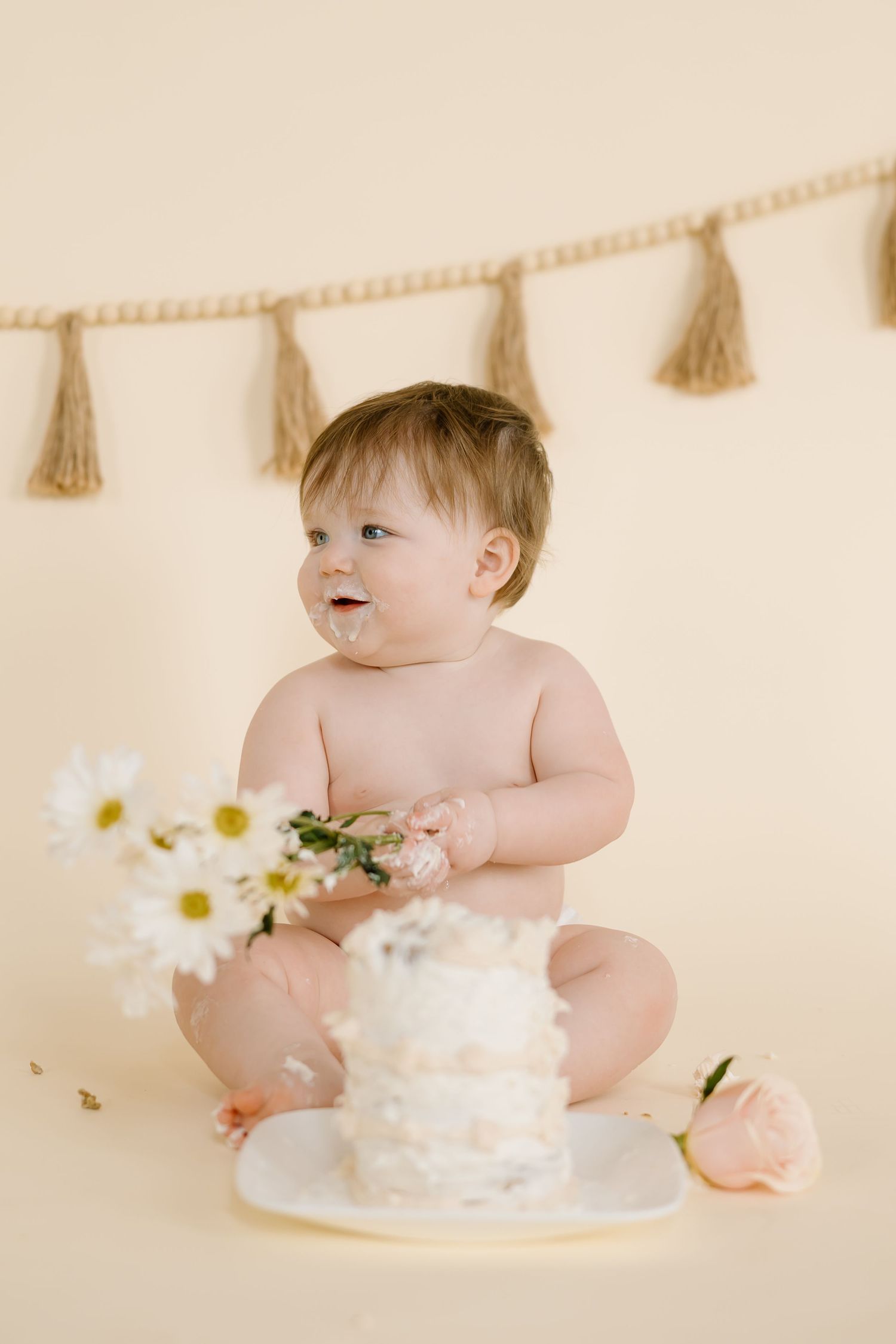 Baby holds flowers next to white cake with tassel garland backdrop during photoshoot.
