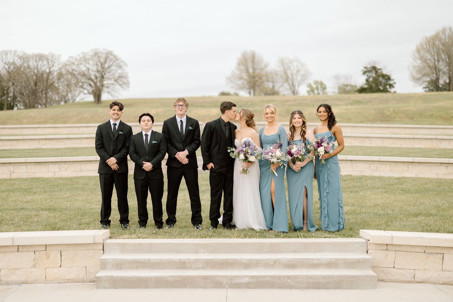 Wedding party in black suits and light blue dresses poses on stone steps outdoors.