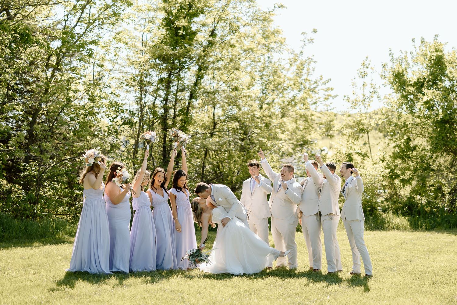 Wedding party strikes fun celebratory pose outdoors with bridesmaids in blue and groomsmen in beige.