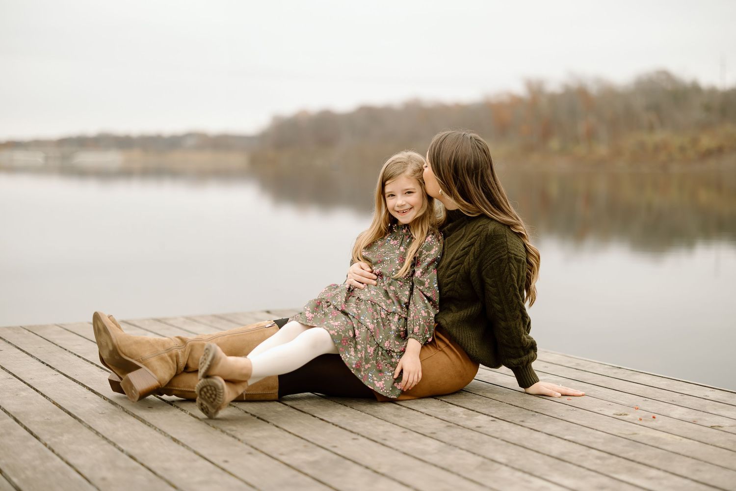 Parent and child sit casually together on wooden dock by calm lake during fall photo session.