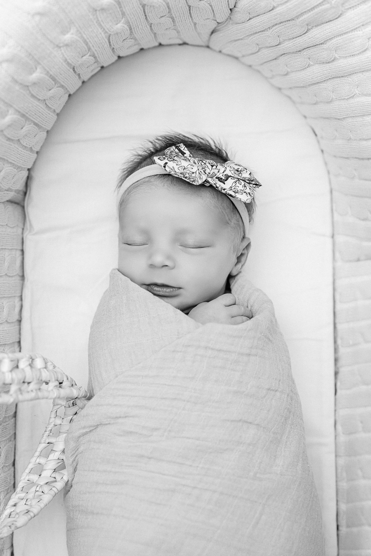 Sleeping newborn wrapped in white swaddle with sparkly bow headband resting in plush bassinet.