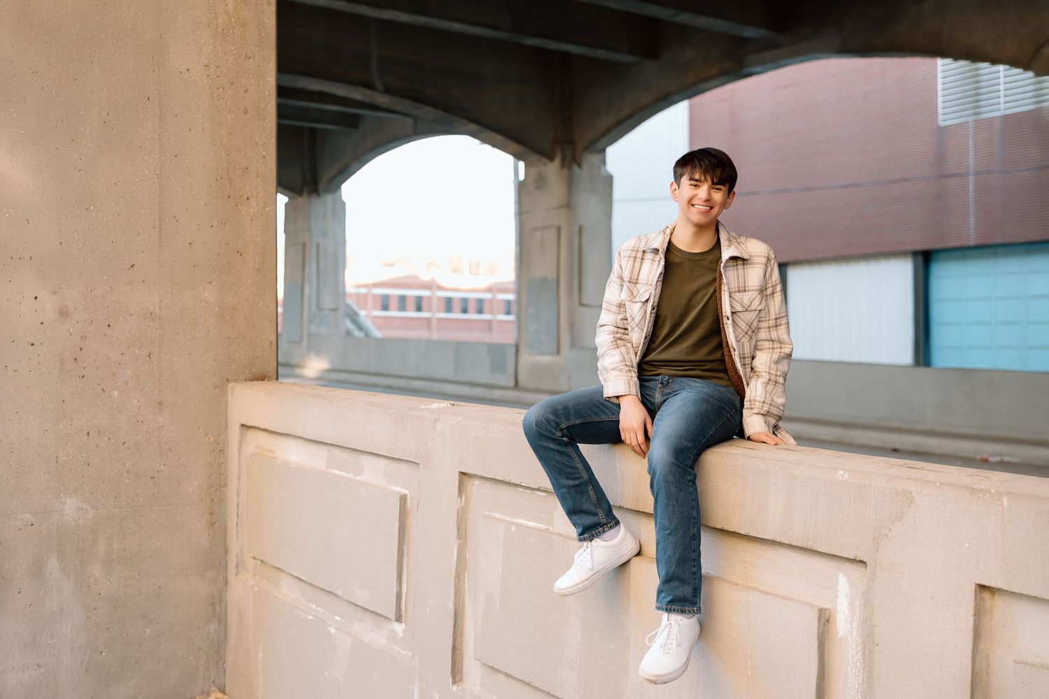 Sitting casually on concrete ledge under bridge wearing plaid jacket and jeans.