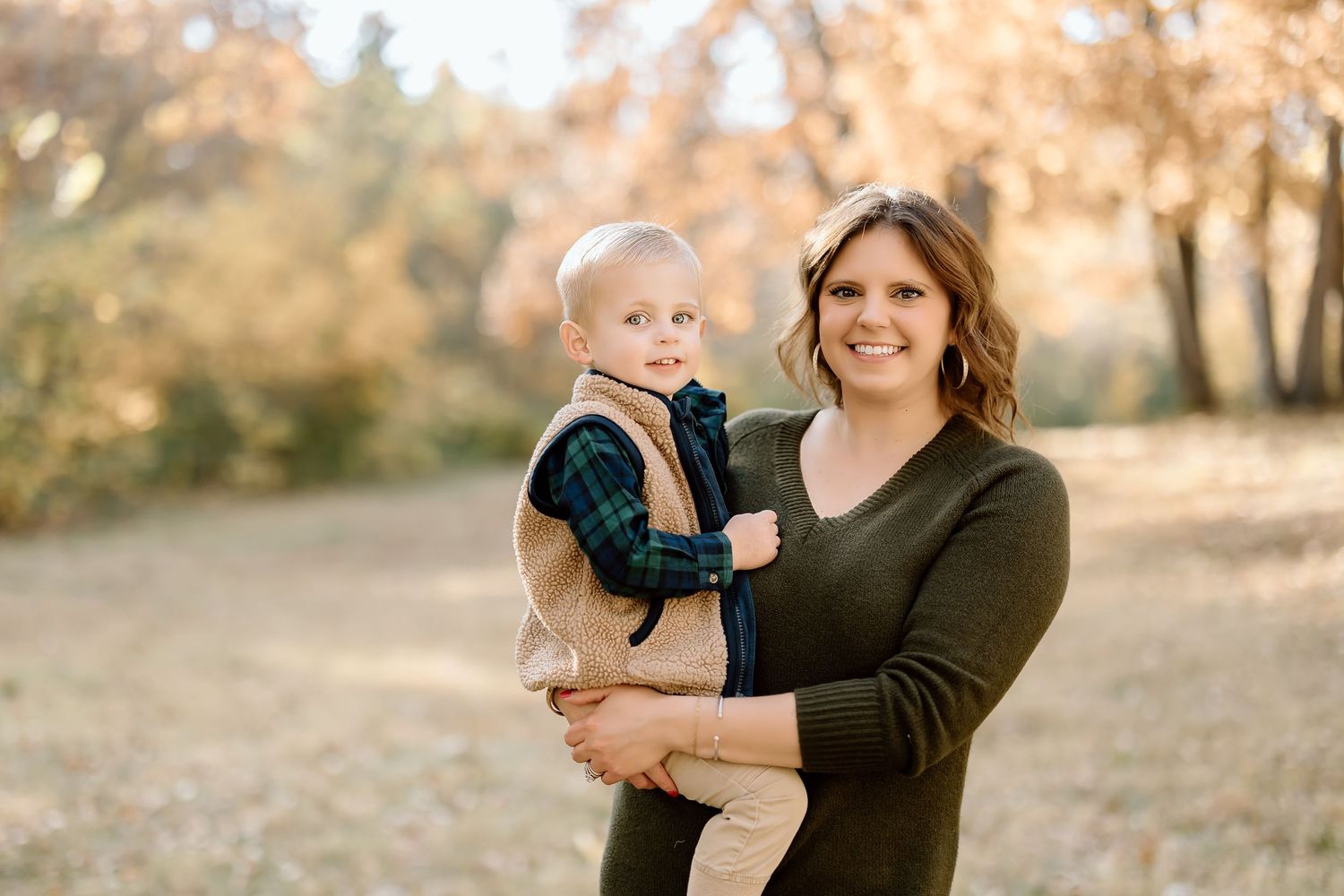 A parent in a dark sweater holds a small child wearing beige and navy in an autumn outdoor setting.