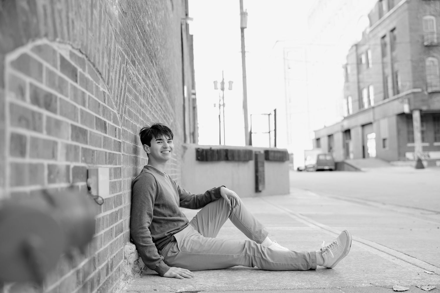 Black and white portrait of person sitting against brick wall in urban environment.