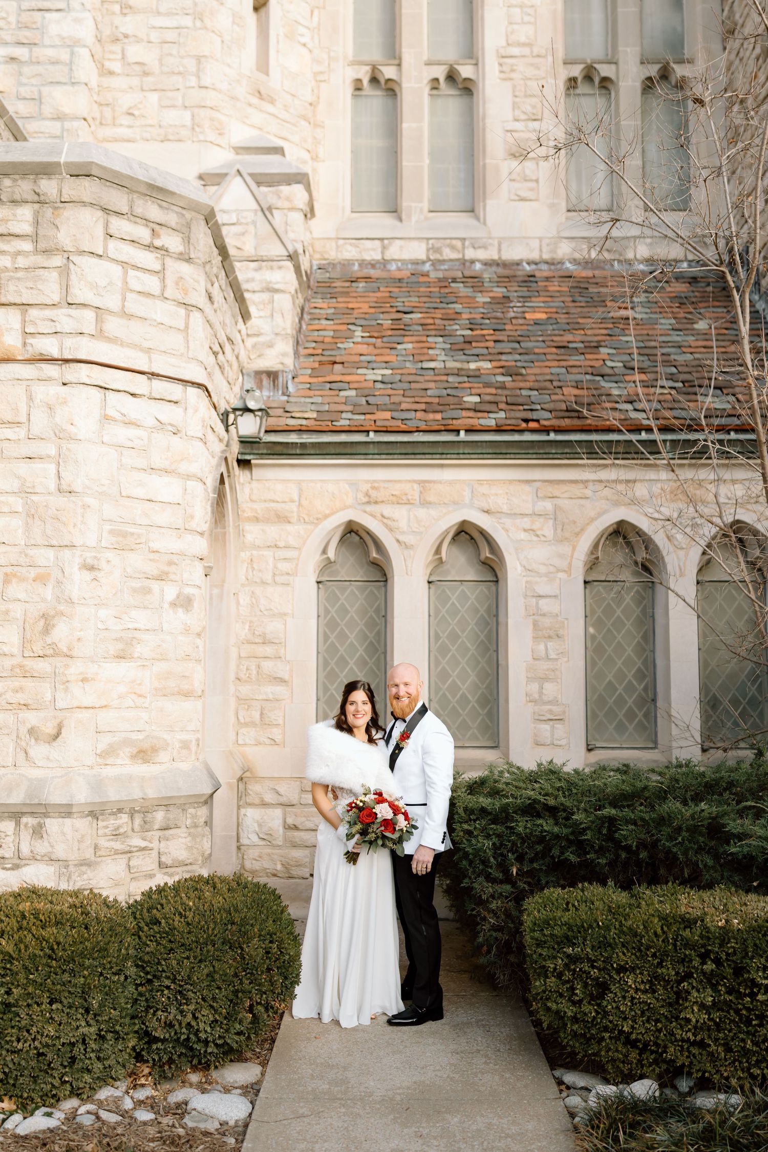 A couple shares a romantic moment outside a gothic stone church with arched windows and manicured hedges.