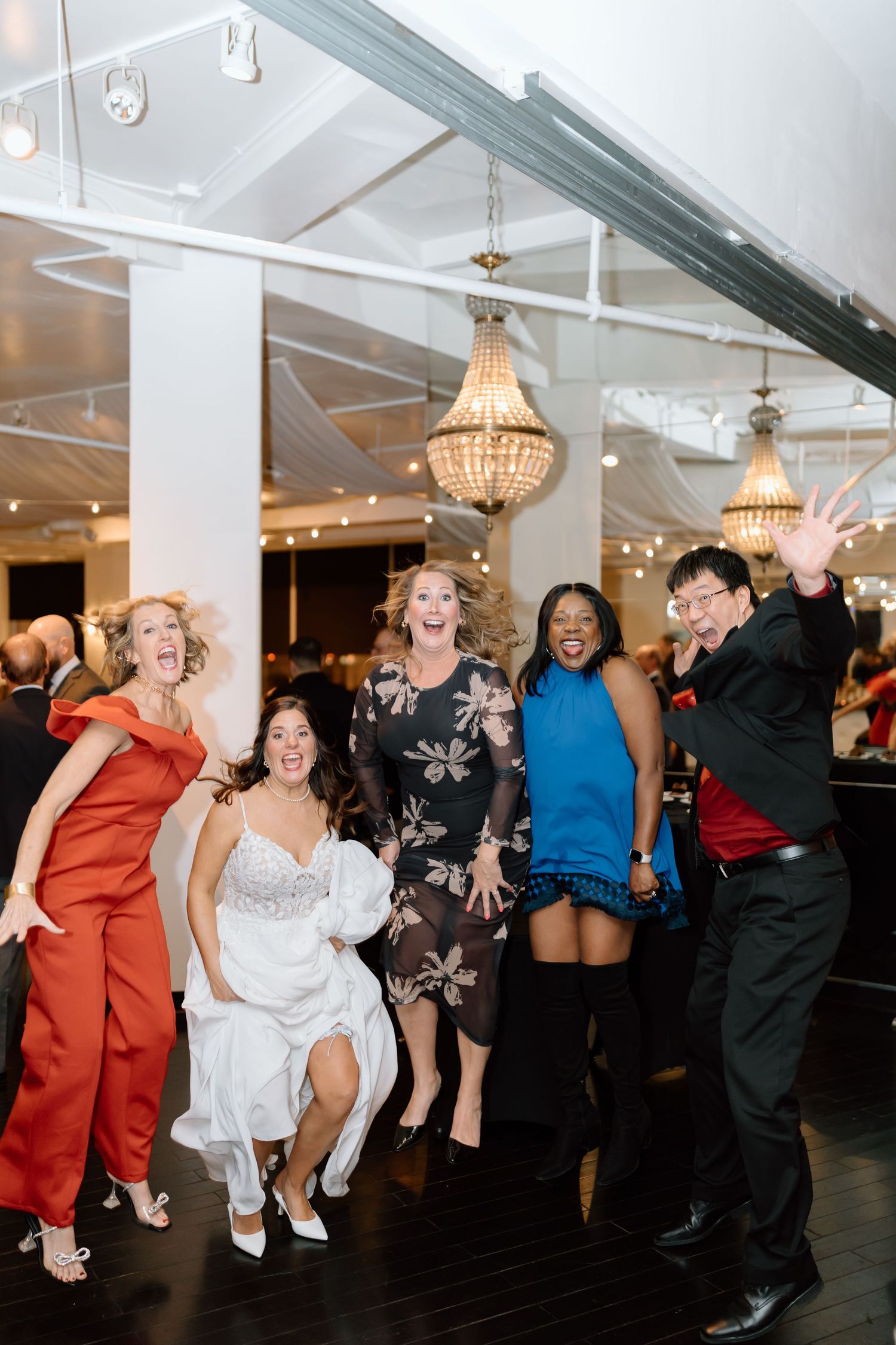 A group of wedding guests dancing and celebrating together on a dark dance floor under crystal chandeliers.