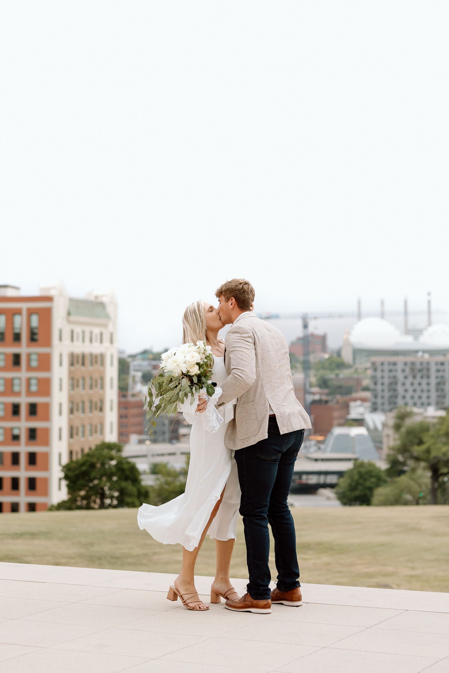 A romantic couple shares a tender moment on a hilltop overlooking a cityscape on a sunny afternoon.