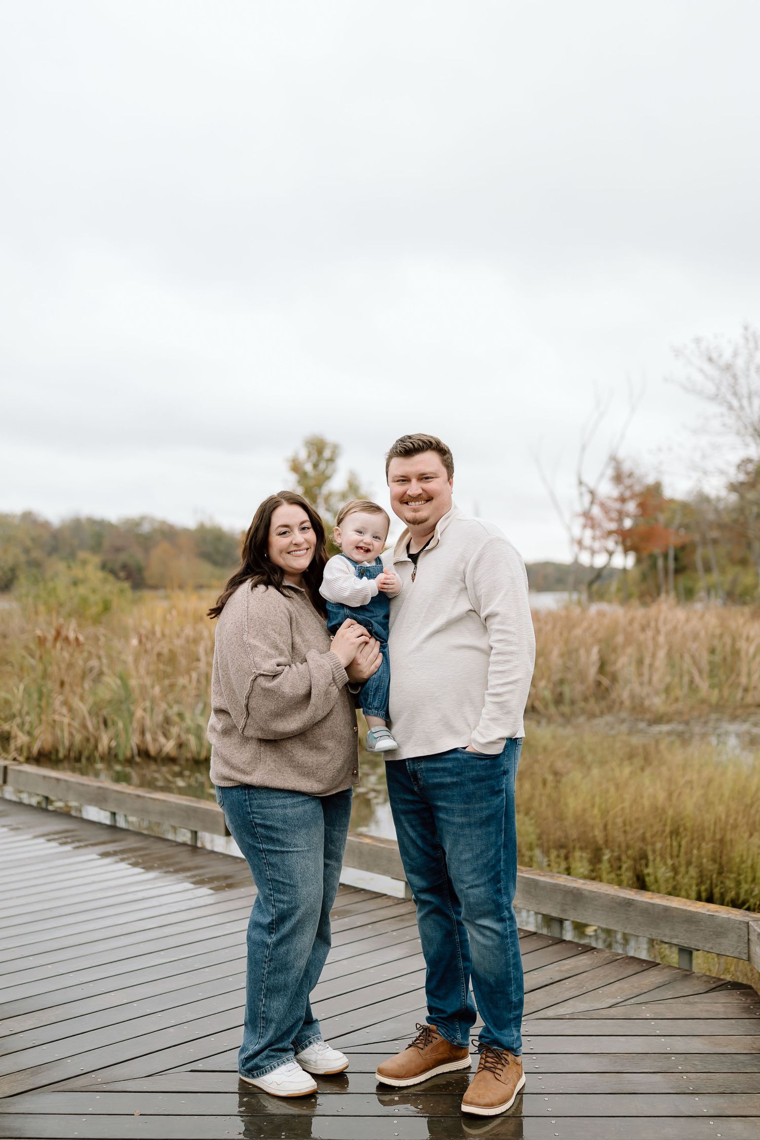 A family stands together on a wooden boardwalk surrounded by autumn marsh grasses on an overcast day.