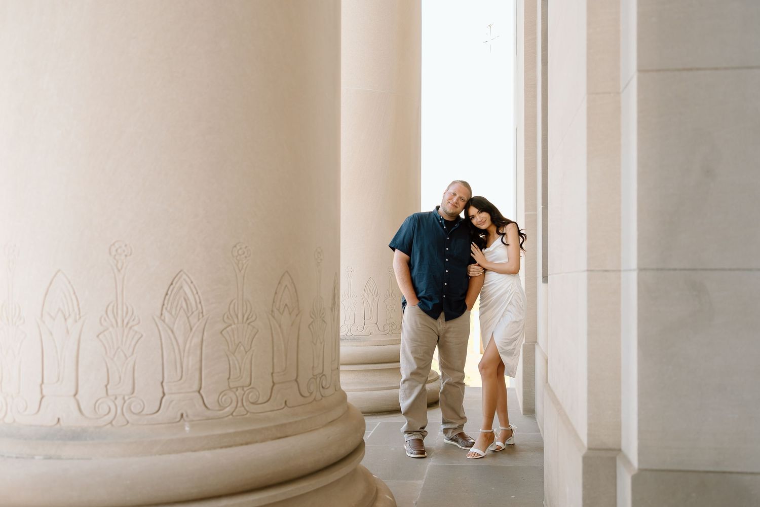 A couple shares a romantic moment between elegant white marble columns in a classical architectural setting.
