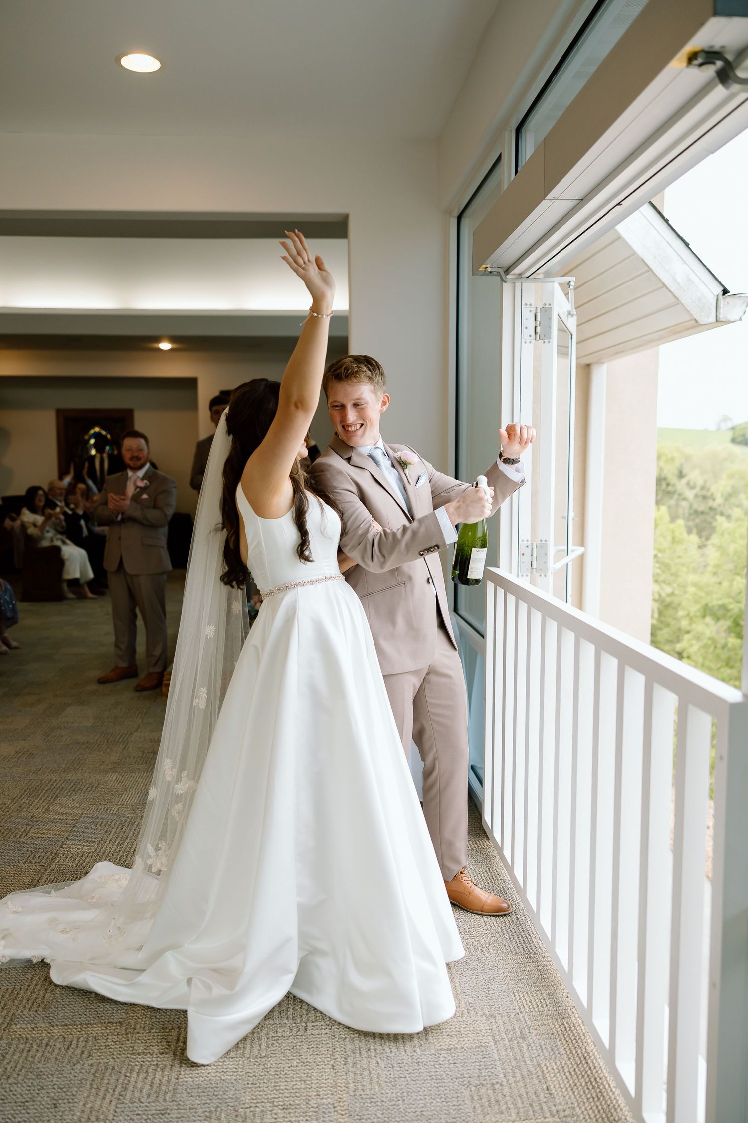 A couple in wedding attire dances playfully together in a bright room with large windows.
