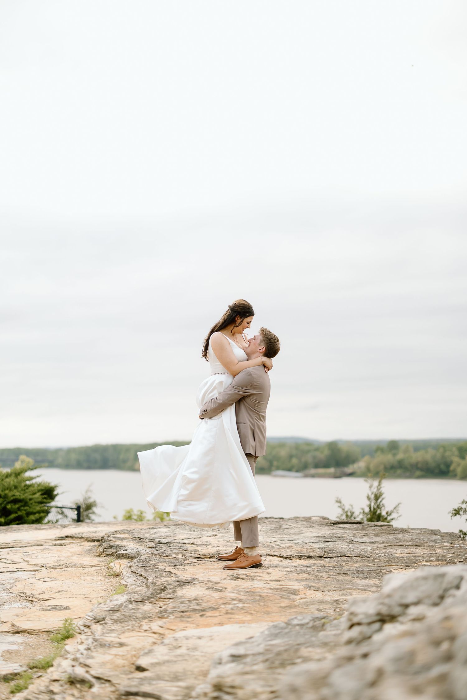A romantic couple embraces on a rocky cliff overlooking a serene lake on their wedding day.