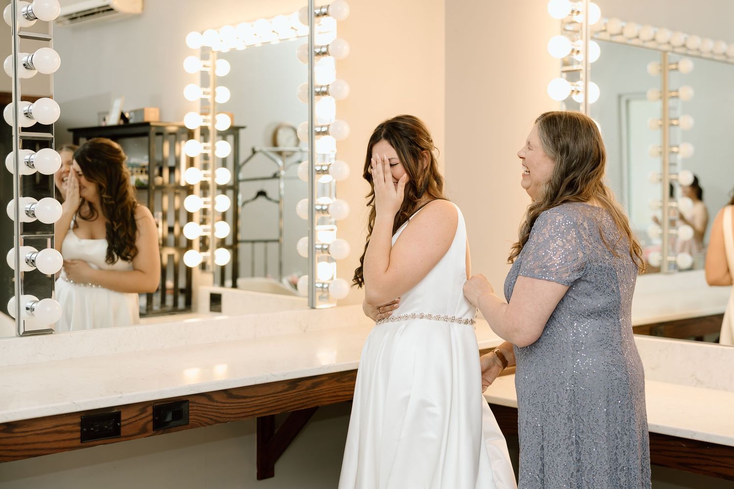A makeup artist works on a client in a brightly lit dressing room with illuminated mirrors.