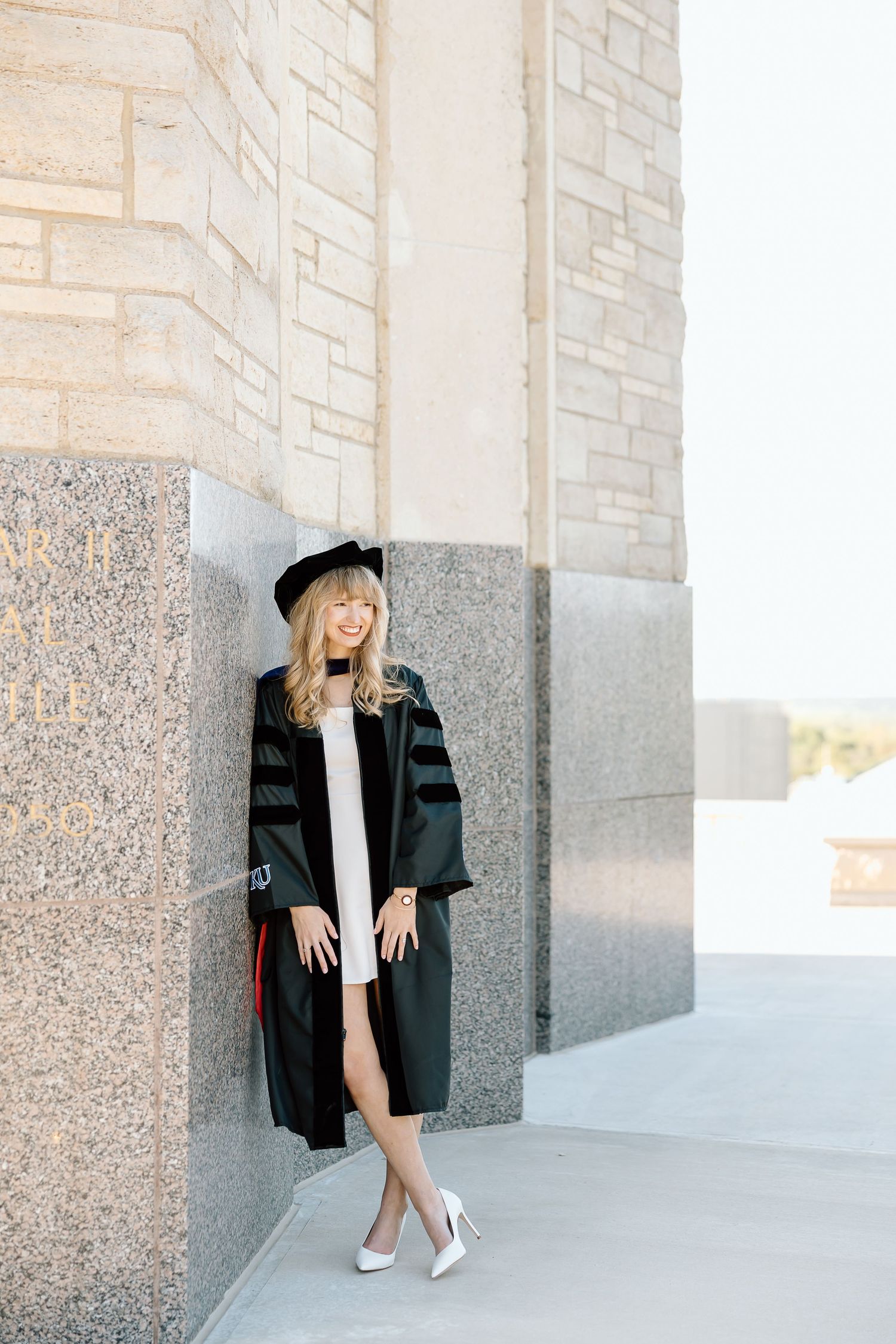 Fashion model in black blazer and white heels poses against stone wall in architectural setting.