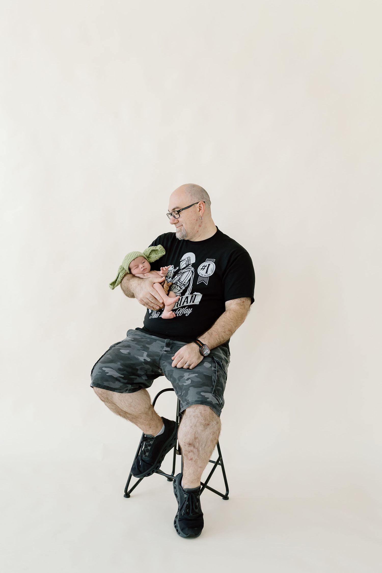 Person wearing black t-shirt and camo shorts sits in various poses on a chair against white background.
