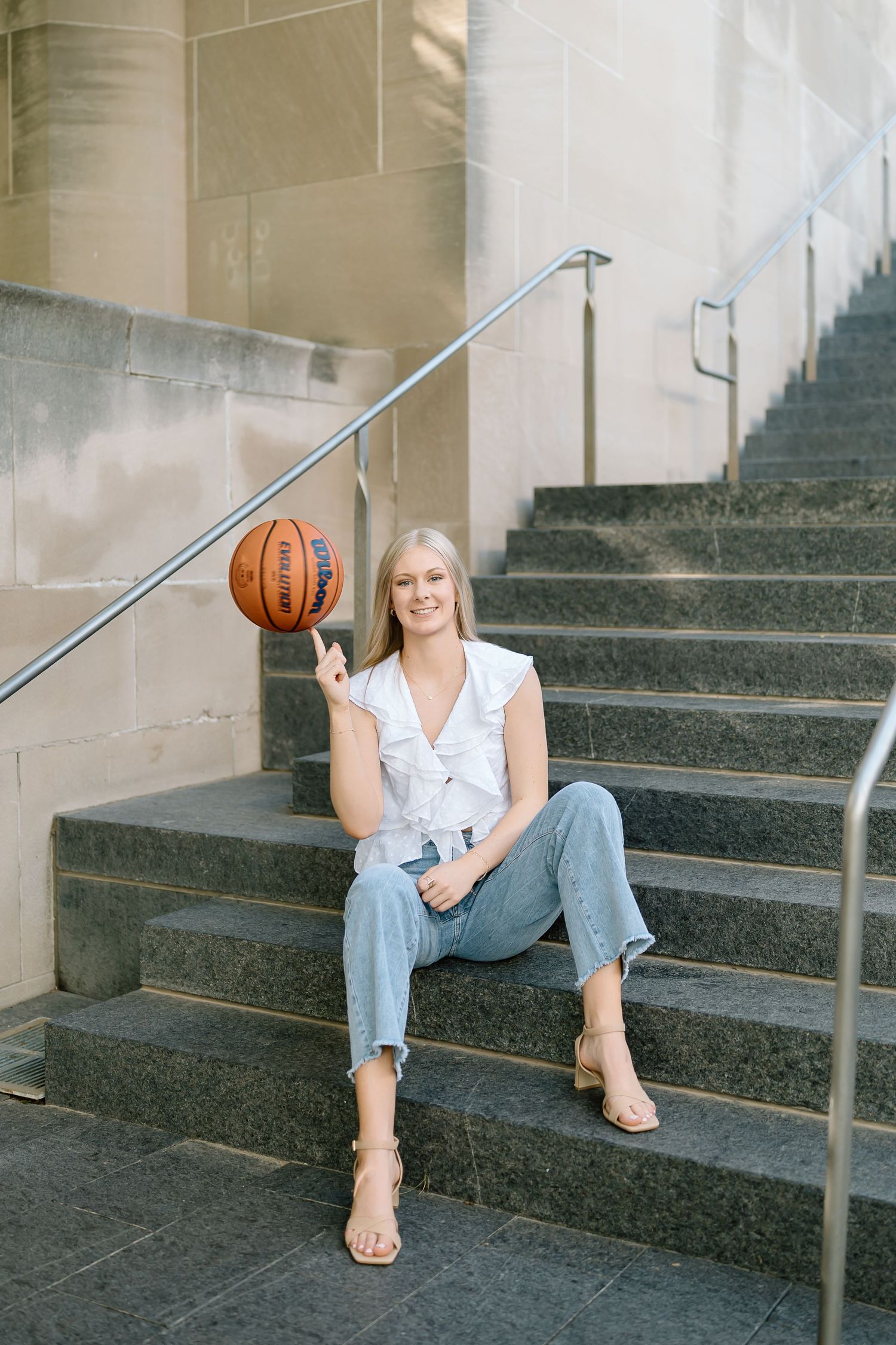 A casual portrait on concrete steps featuring a basketball, light ruffled top, jeans and strappy sandals.