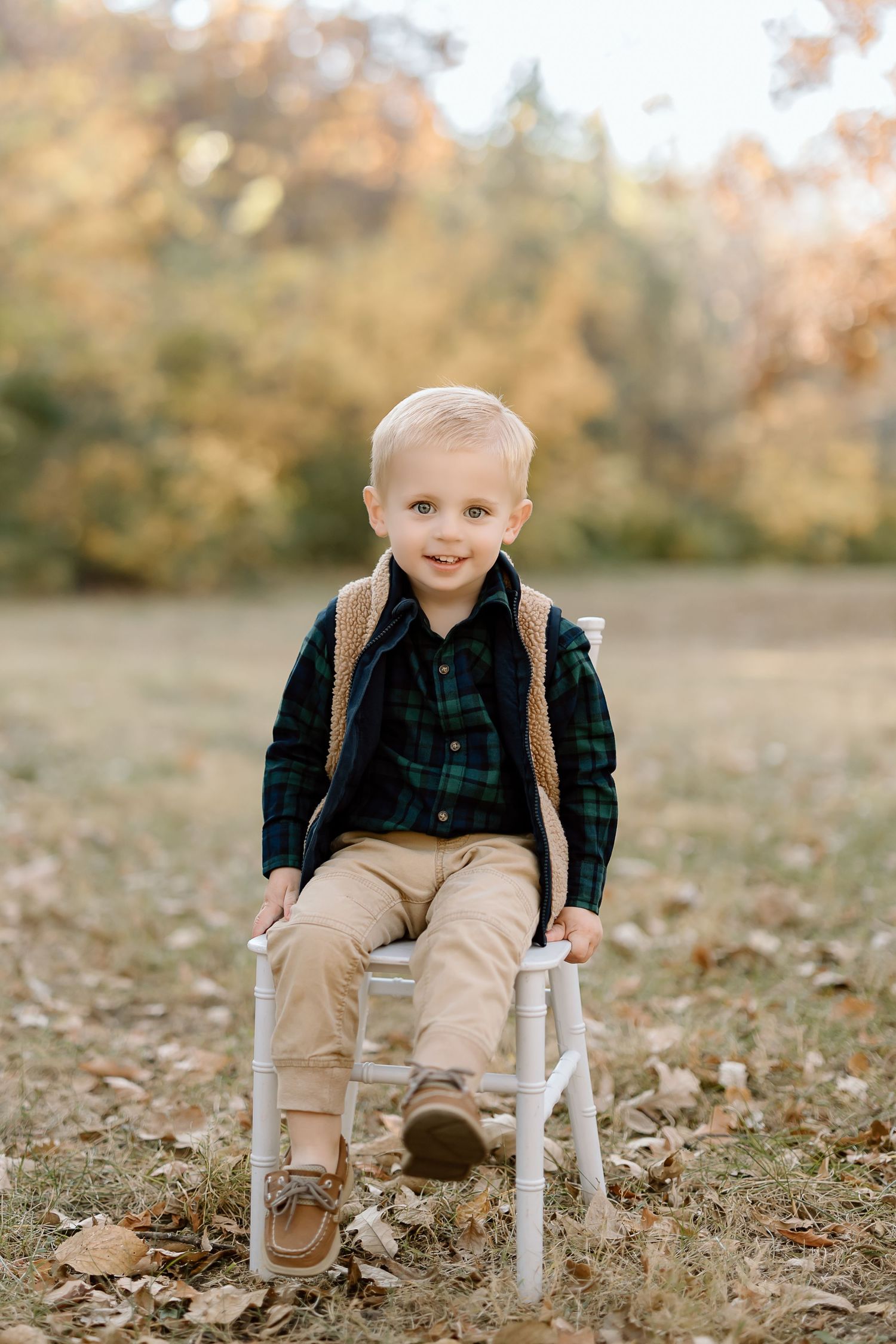 Child in green plaid shirt and vest sits on white chair outdoors during fall.