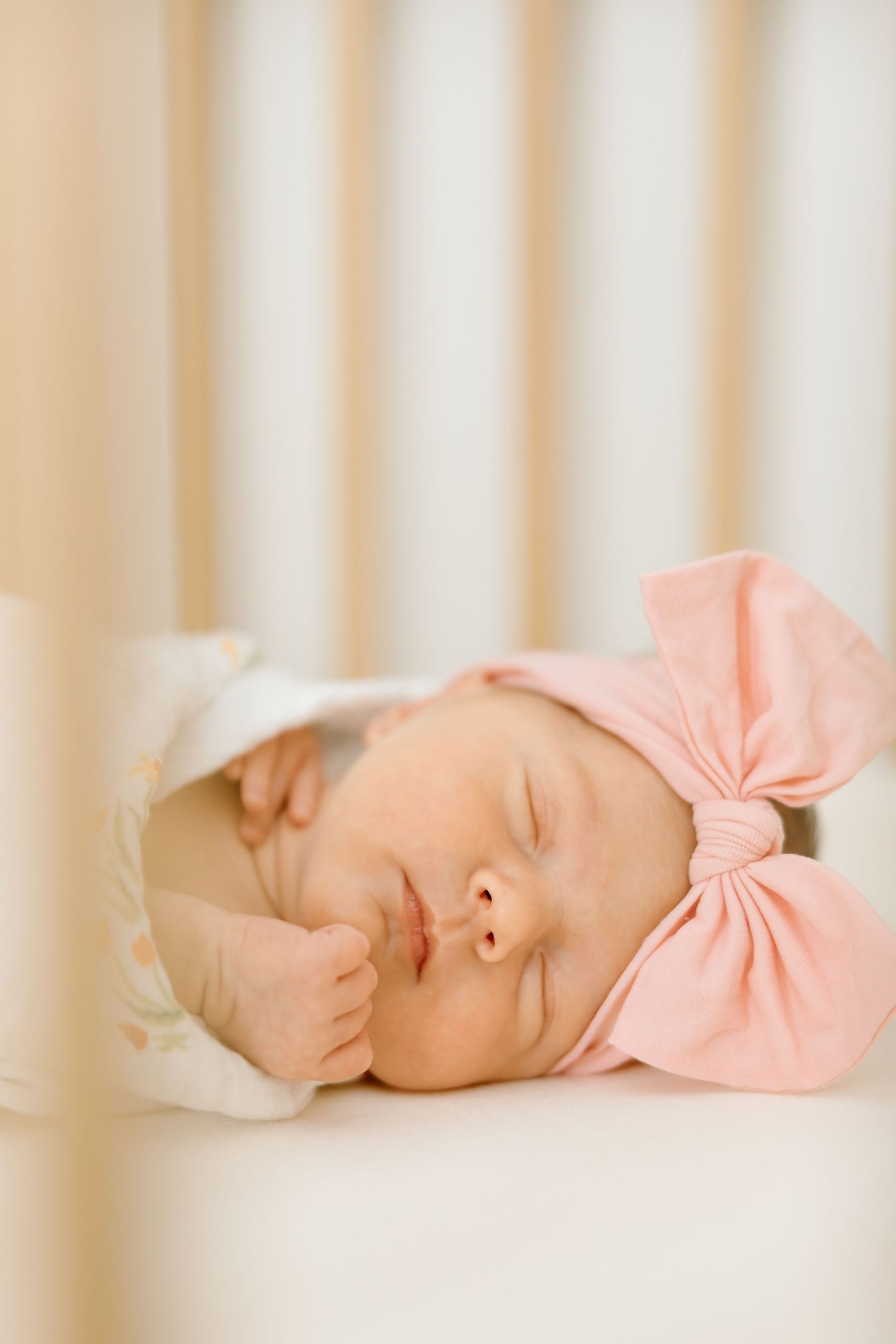 A sleeping baby wearing a pink bow headband rests peacefully in a white crib.