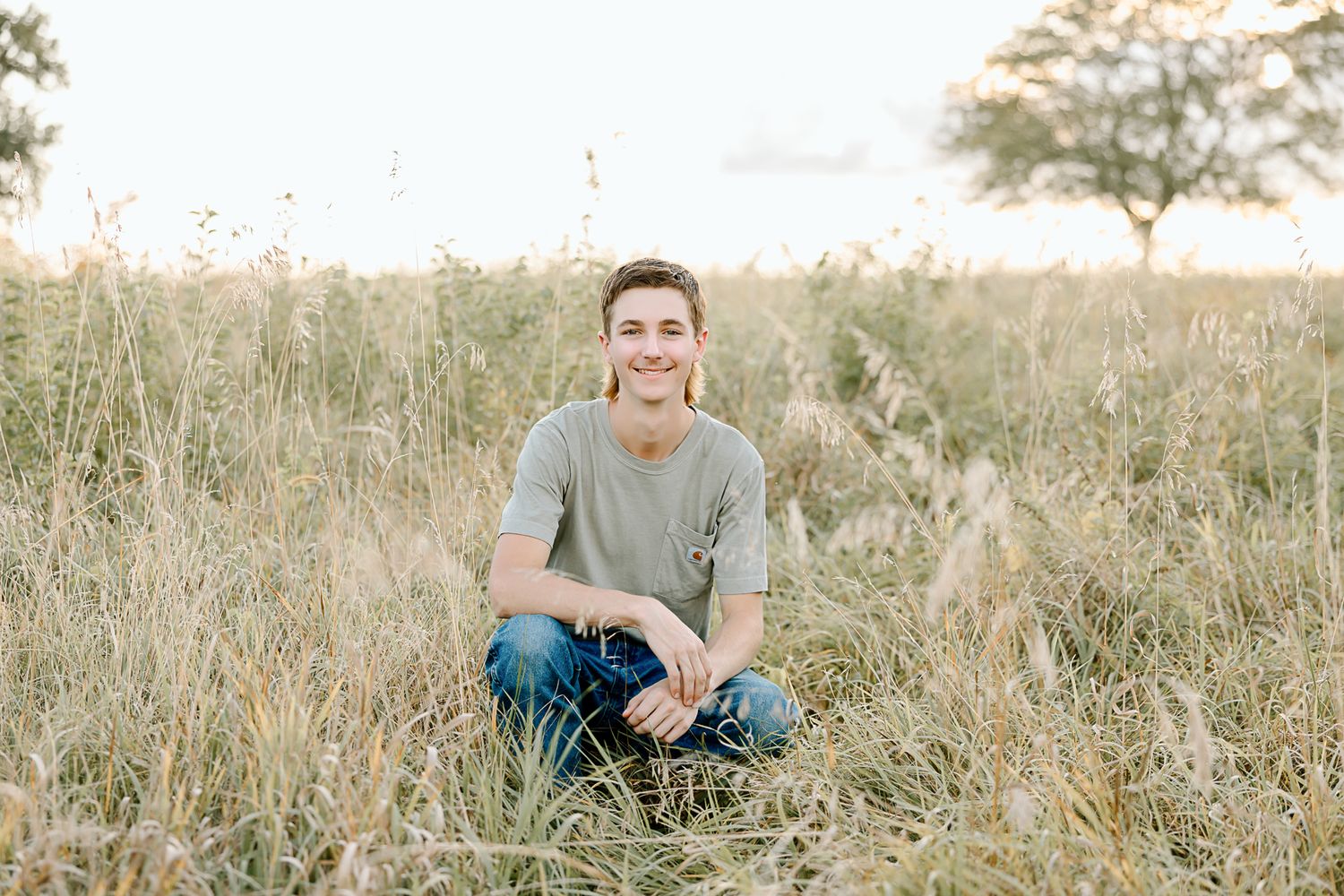Person sitting cross-legged in a sunlit field wearing blue jeans and gray shirt.