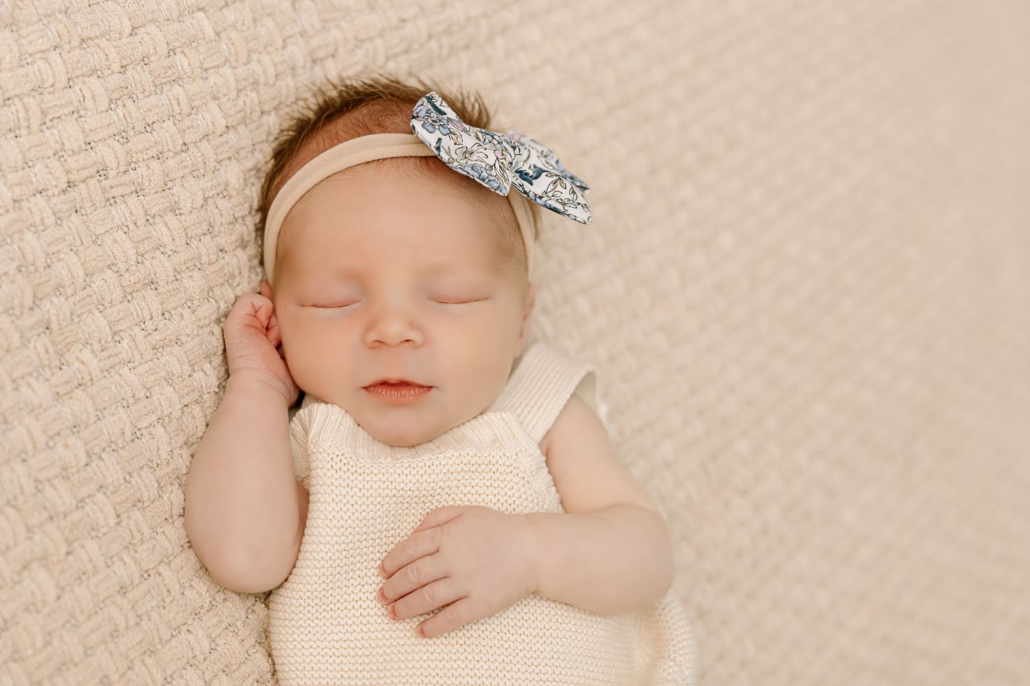 Sleeping newborn wearing cream outfit and sparkly bow headband on textured beige blanket.