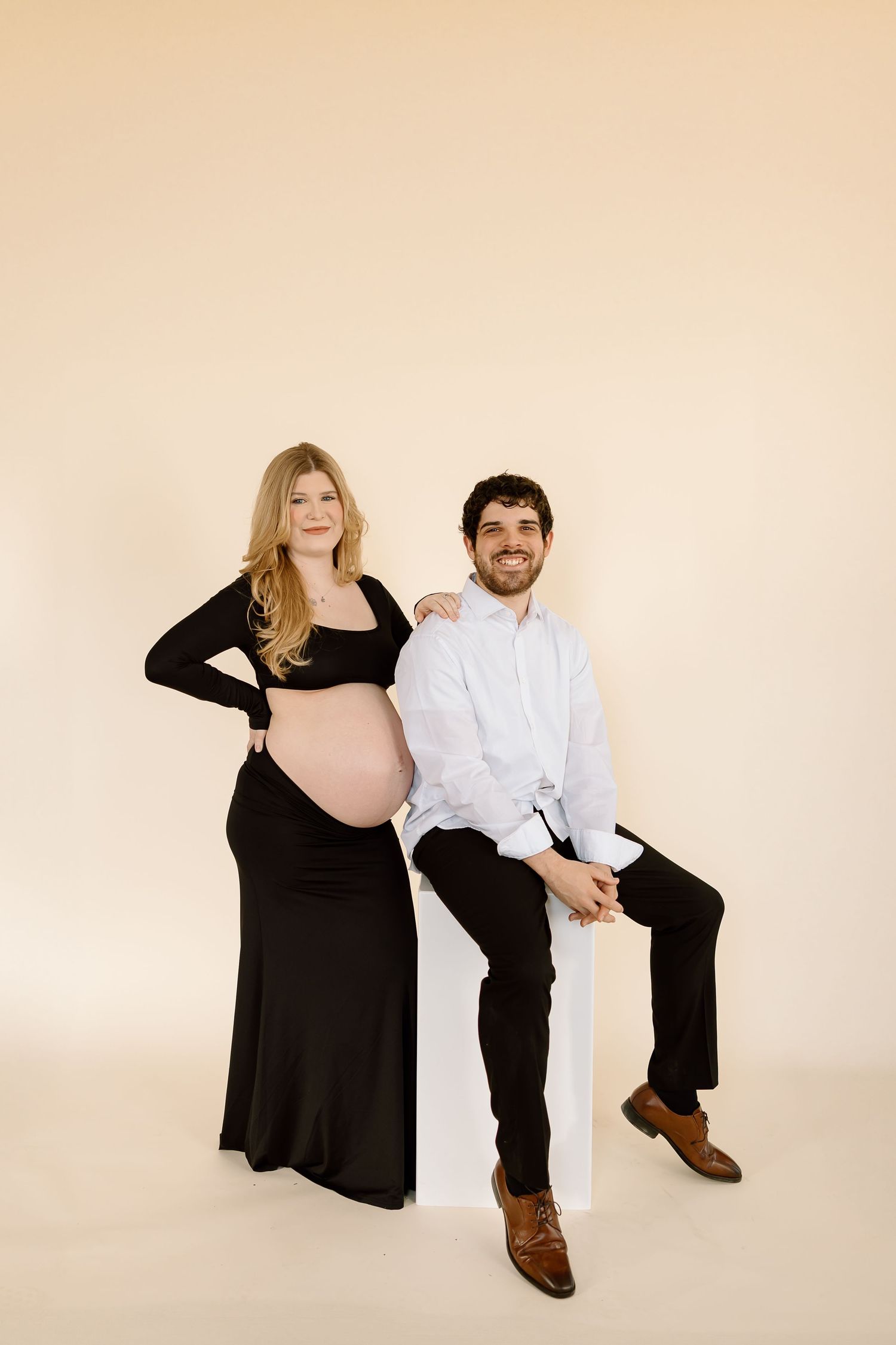 Expecting couple poses together on white block against beige backdrop.