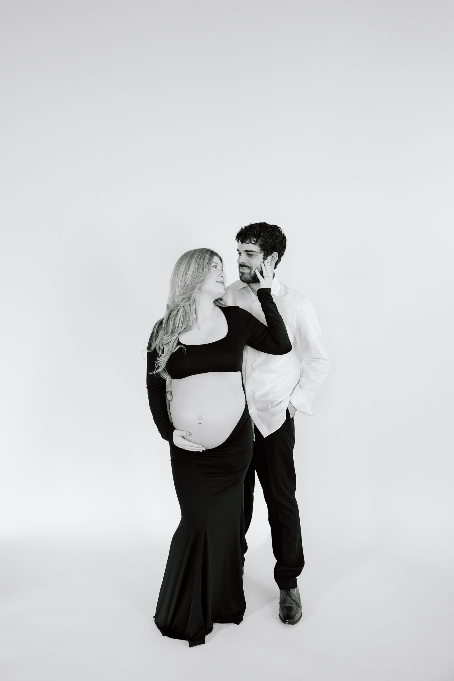 Black and white maternity portrait of a couple in formal wear embracing against a light backdrop.