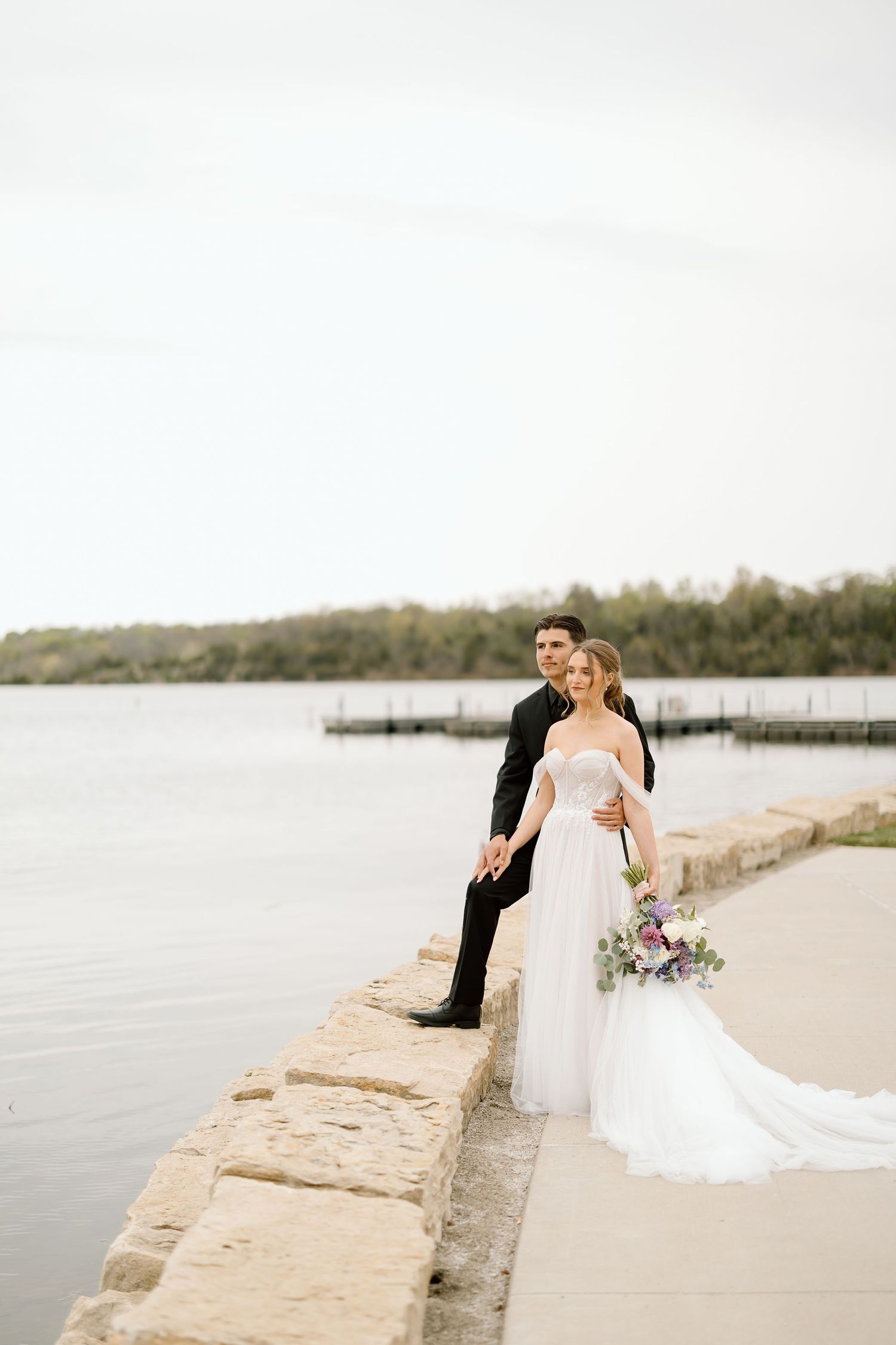 Bride with bouquet and groom sit on stone wall overlooking peaceful lake.