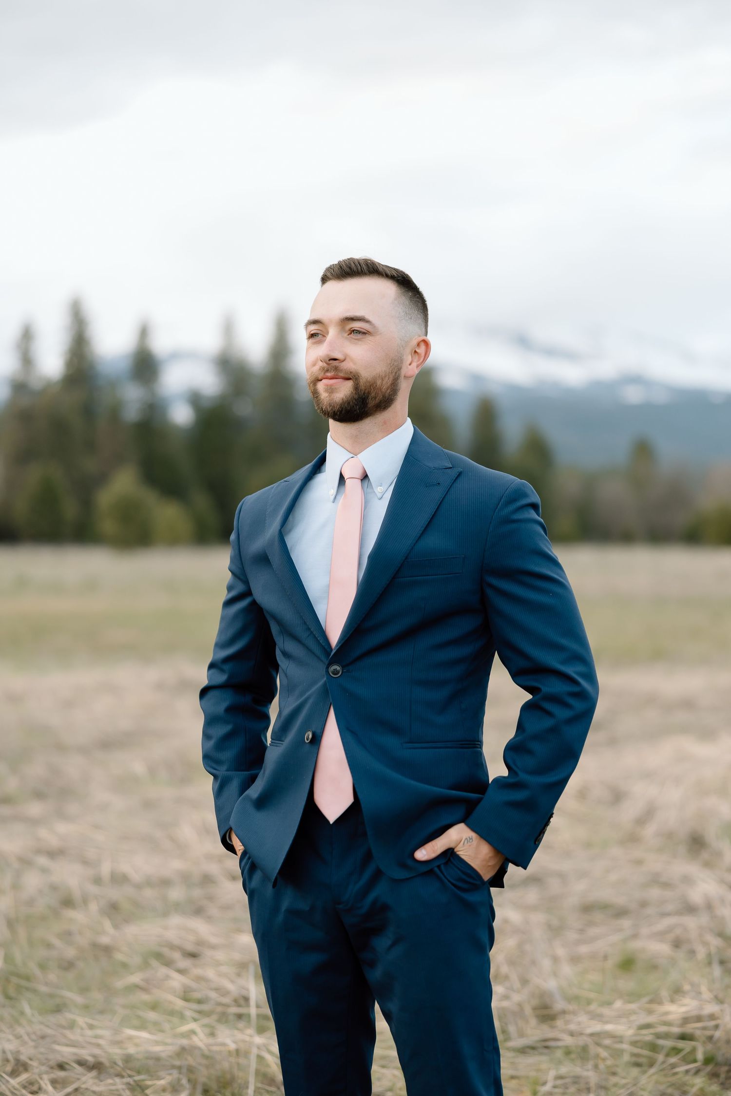 Groom in navy suit with pink tie stands confidently against mountain landscape.
